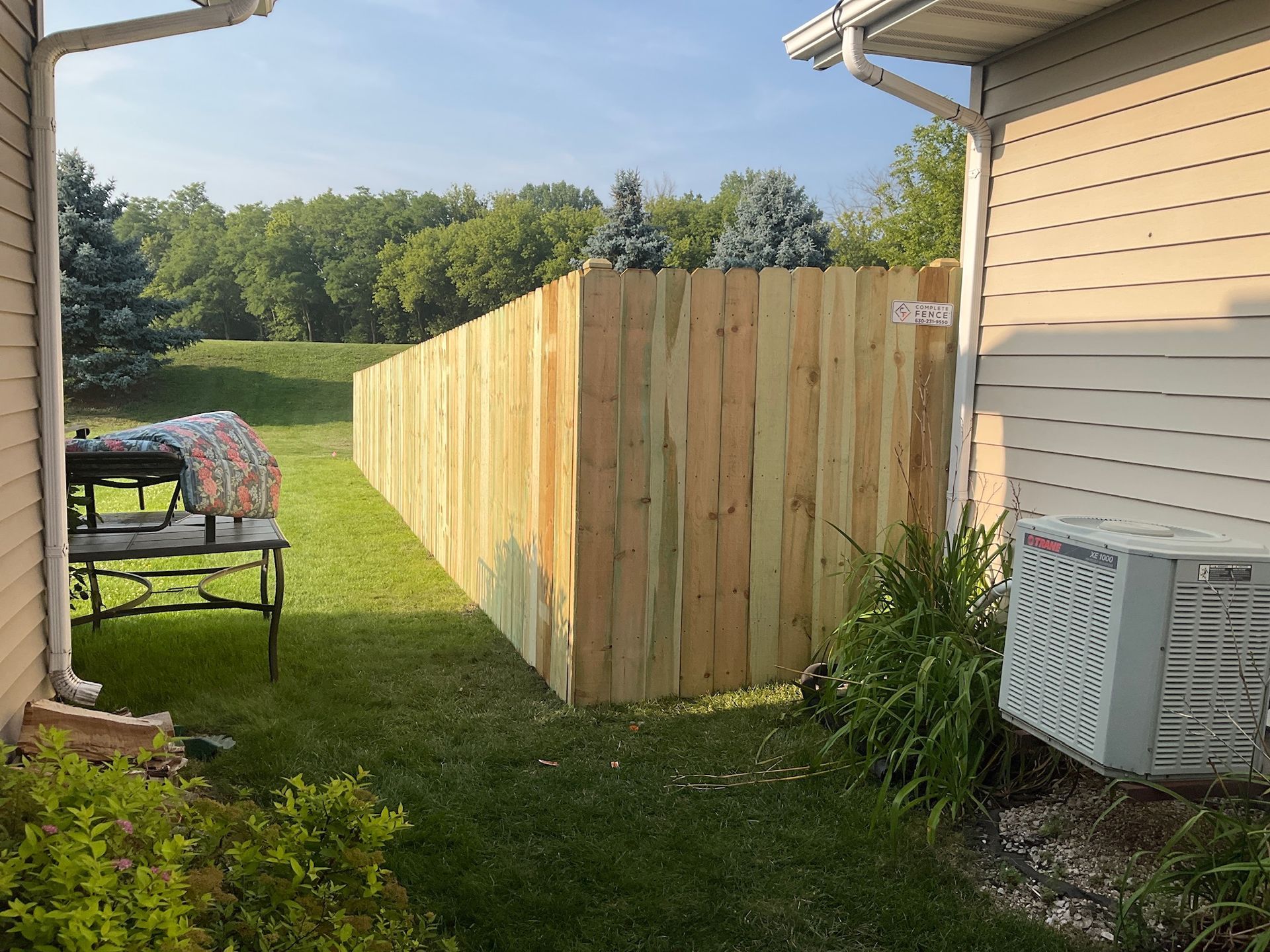 Wooden fence in backyard, separating two houses. Green grass and trees visible in background.