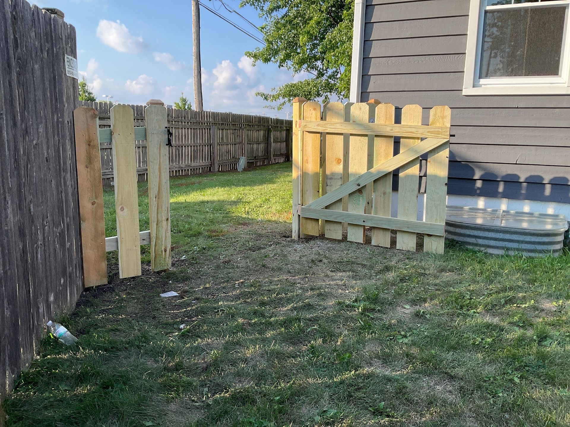 Wooden gate in a grassy yard, opening to a weathered fence and a building with a window.