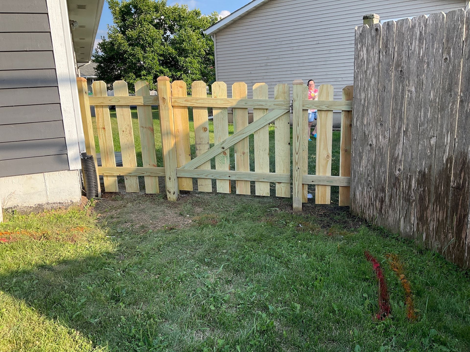 Wooden gate in a grassy yard, connecting a house and a weathered fence. Sunny day.