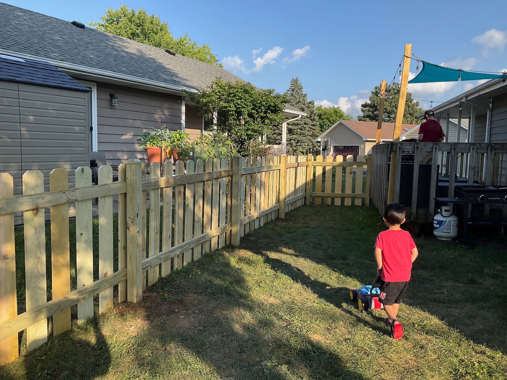A child in a red shirt walks on grass toward a wooden fence. Houses and trees are in the background.