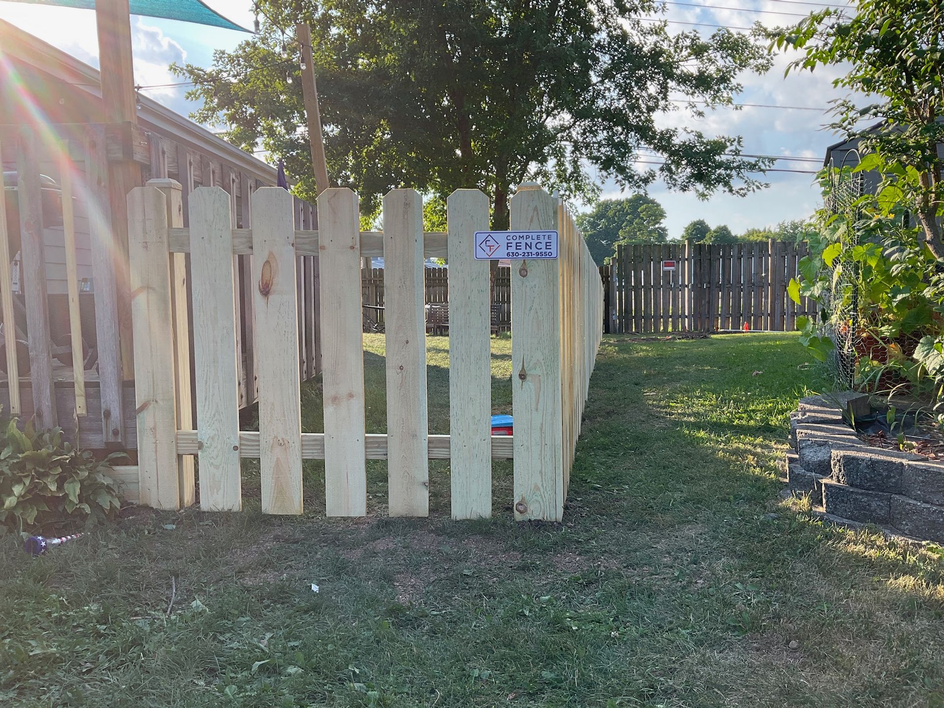 Wooden picket fence in a grassy yard, sunlight, with a sign. Another fence visible in the background.