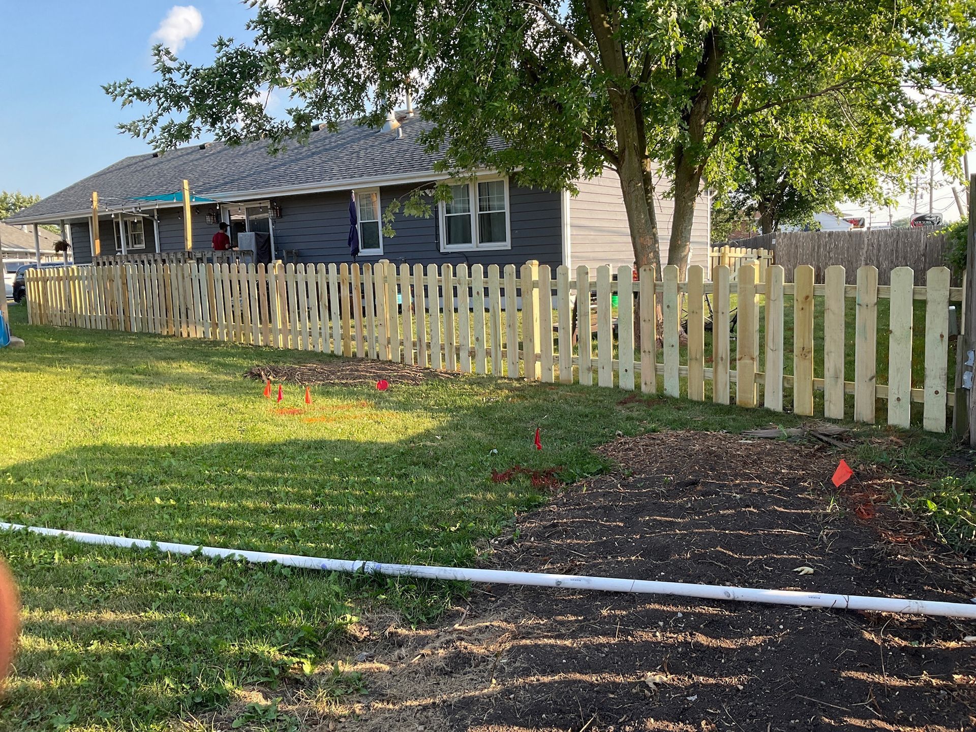 Wooden picket fence in a yard, beside a dark-colored building and a small garden with red flags.