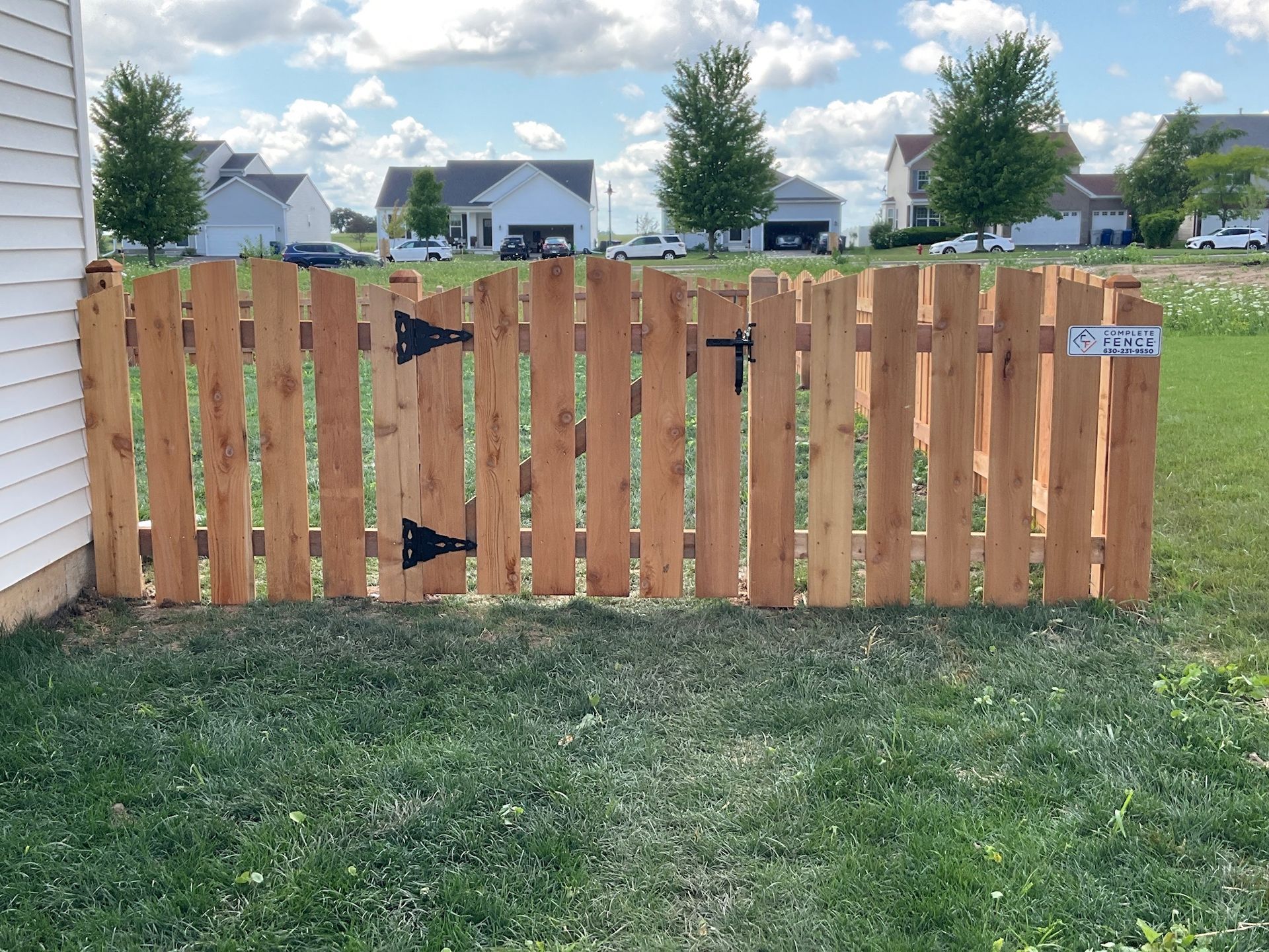 Wooden picket fence with a gate in a grassy yard; houses in the background.