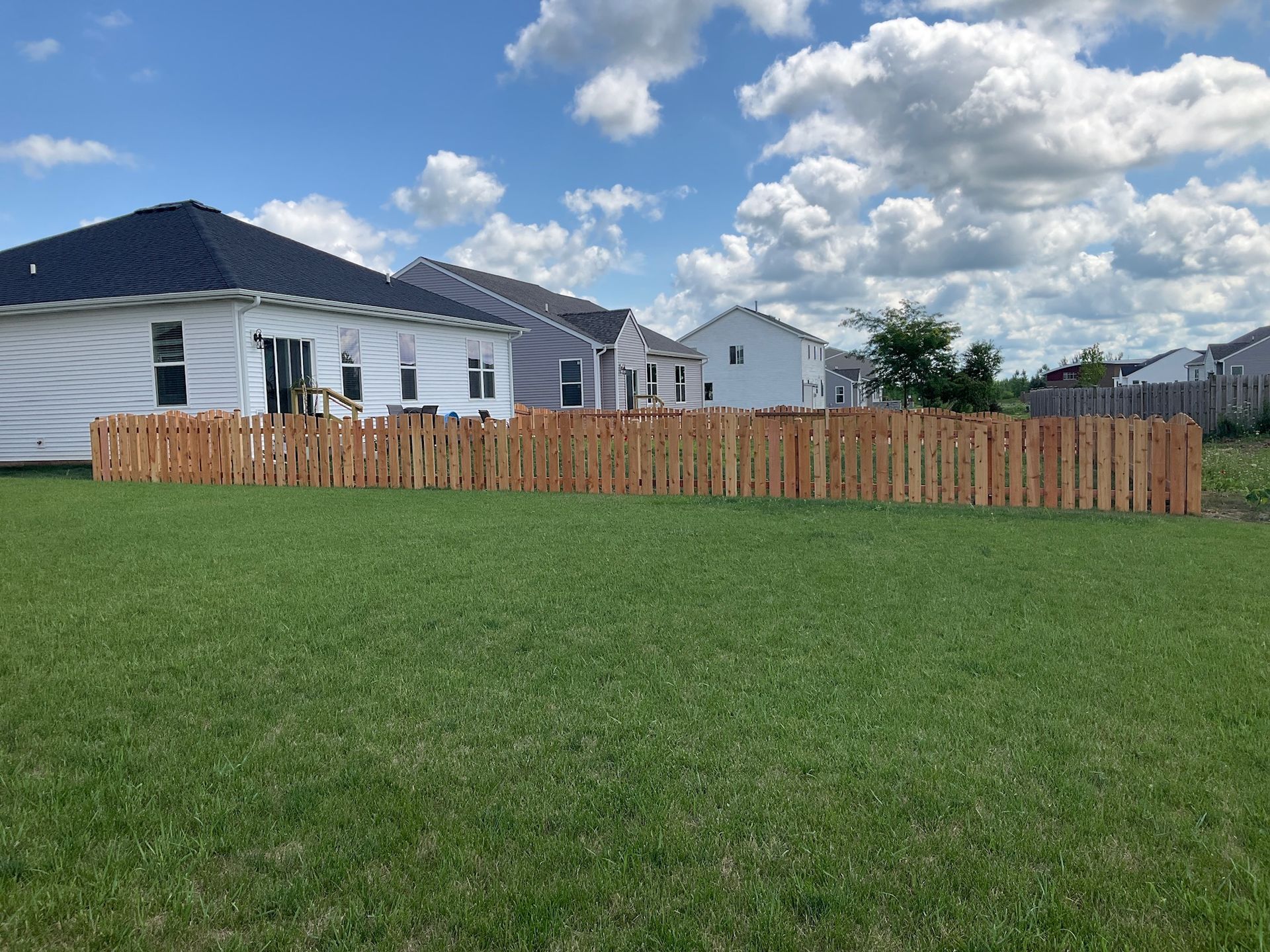 A newly built wooden fence encloses a green backyard, with houses and blue sky in the background.
