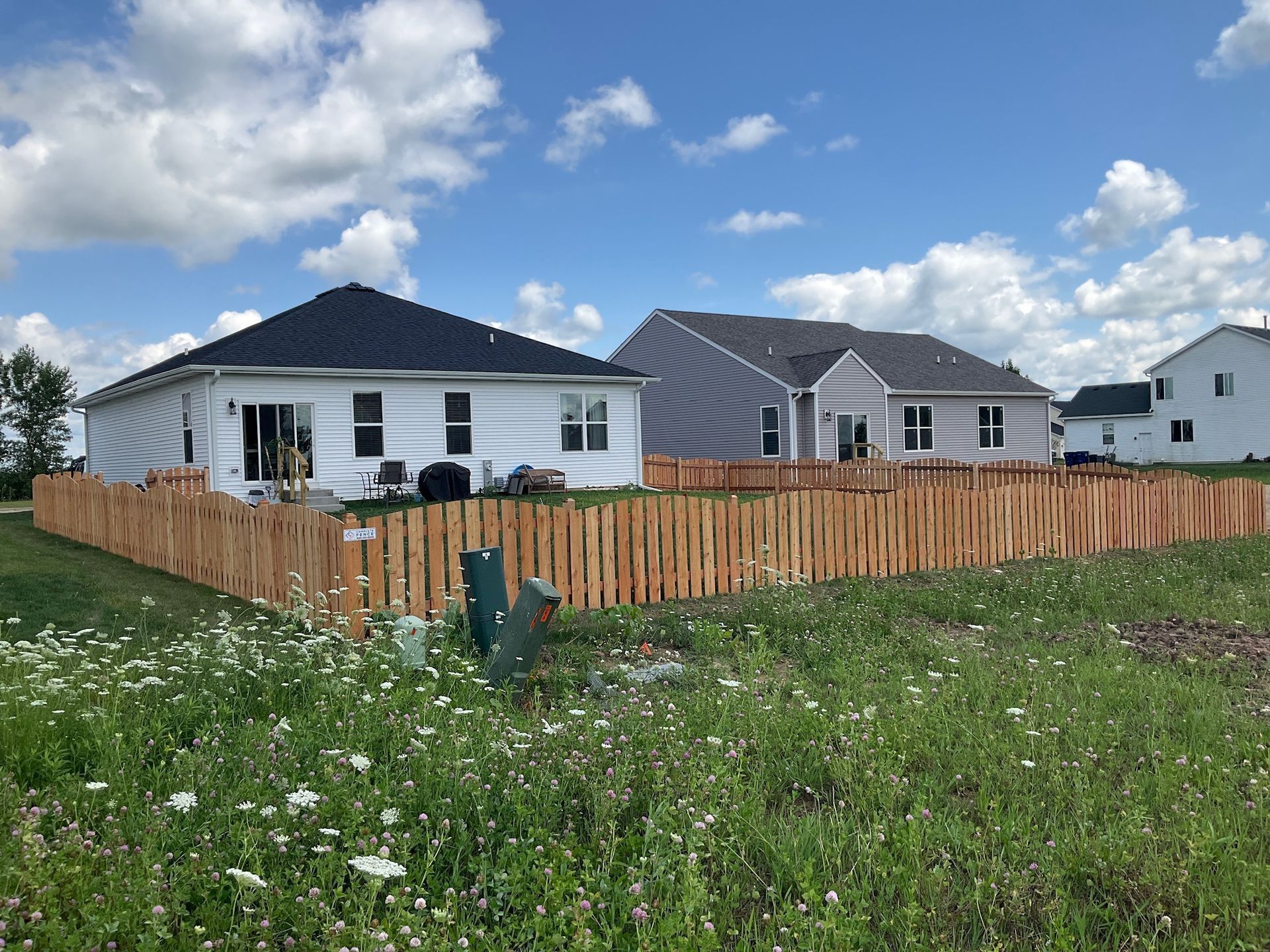 Two houses with a wooden fence in a grassy yard under a blue sky with clouds.