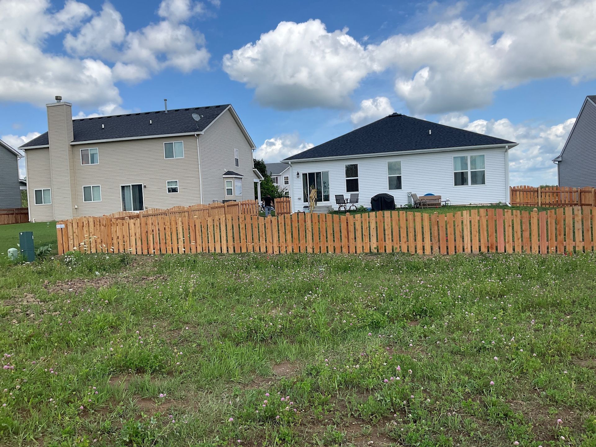 Wooden fence in front of two houses on a grassy lawn under a partly cloudy sky.