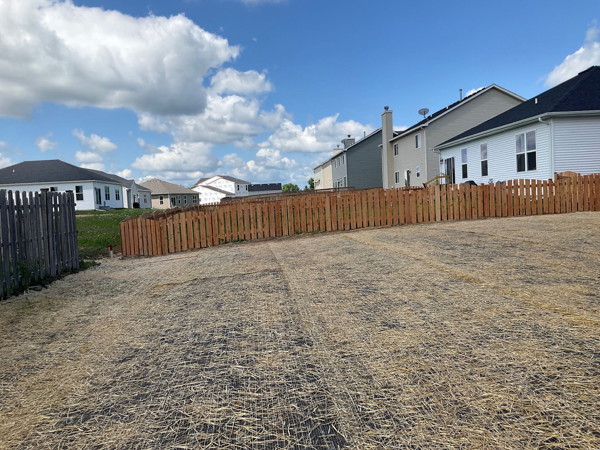 A gravel-covered hillside with a wooden fence and houses against a cloudy sky.