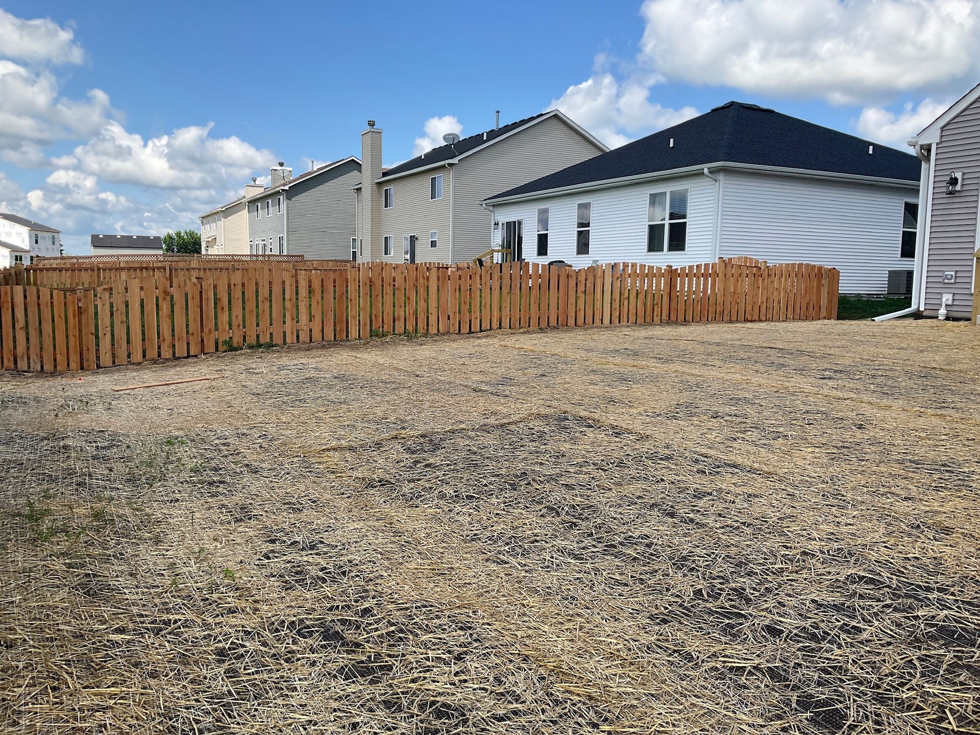Empty gravel lot with a new wooden fence, houses in the background under a blue sky with clouds.