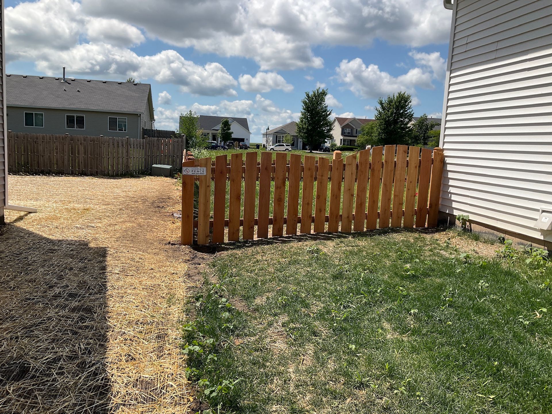 Backyard with grass, wooden fence, and gravel path. Sunny day with a partly cloudy sky.