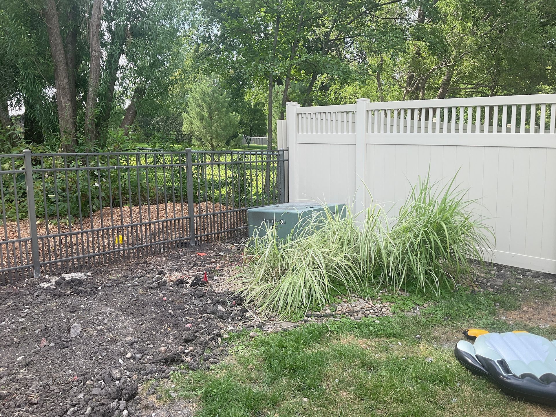 Fenced yard with a green utility box, dry earth, and ornamental grass.