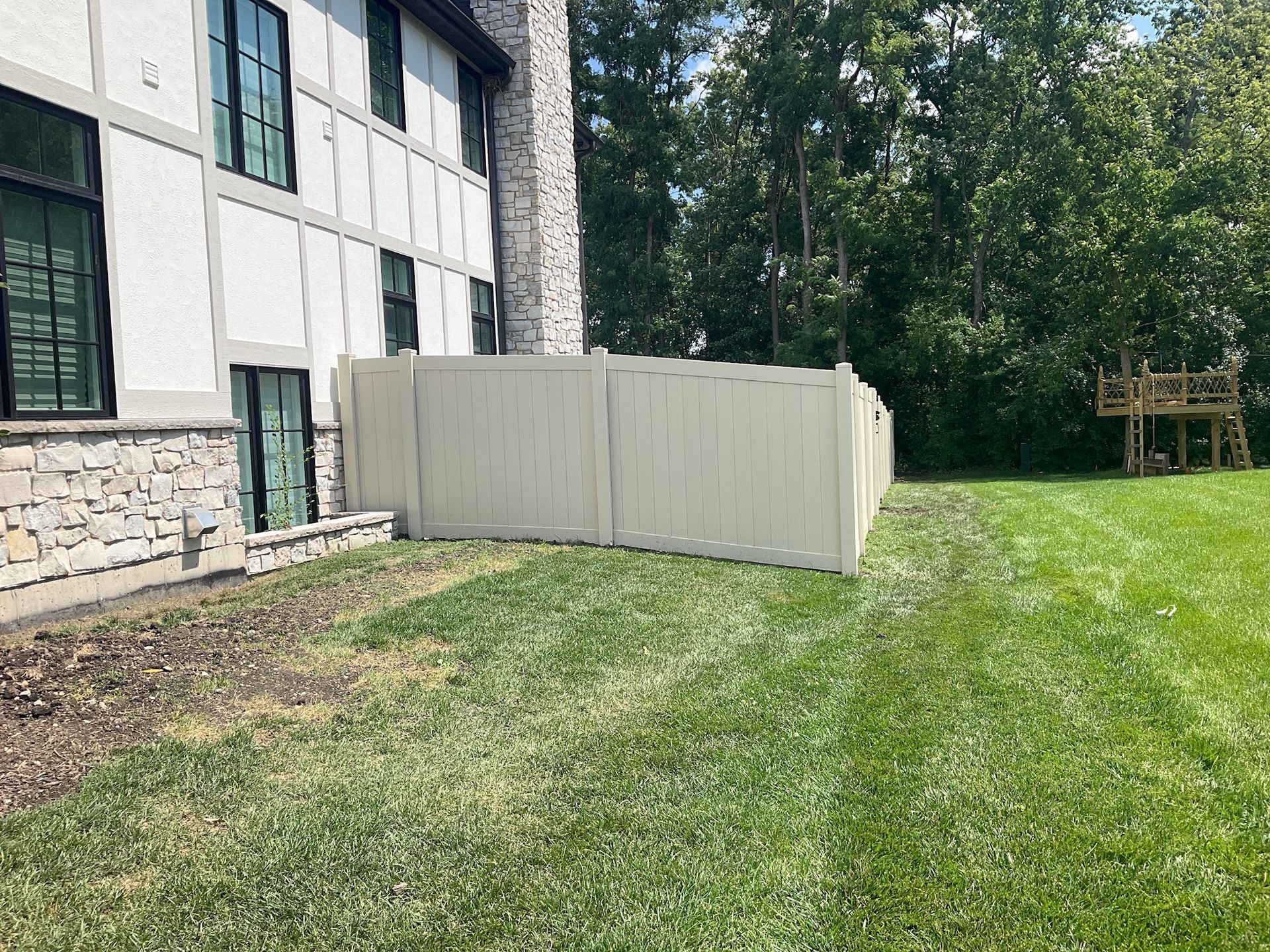A beige fence surrounds a backyard with green grass and a house. A wooden play structure is visible in the distance.