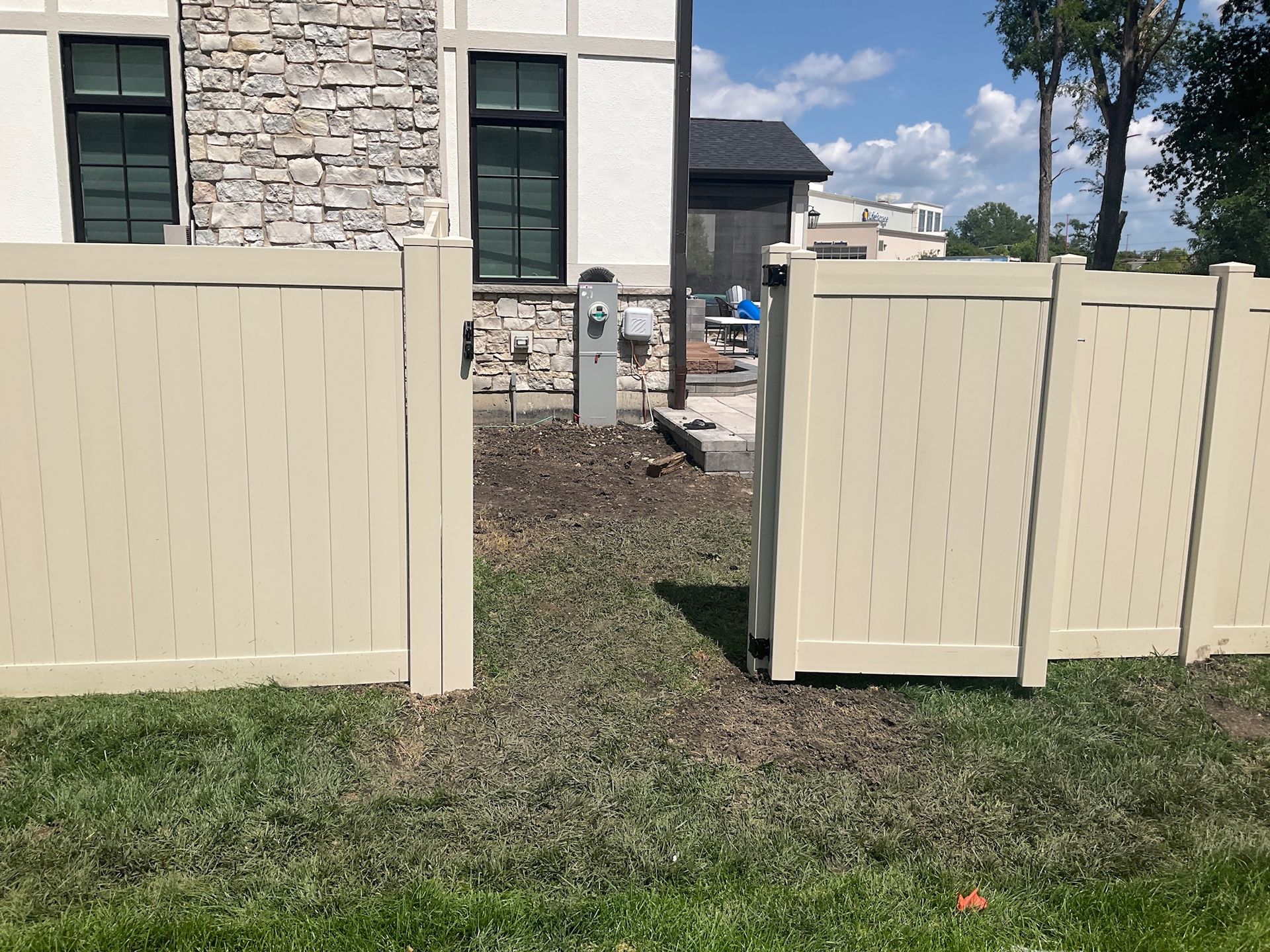 Beige vinyl fence gate open, leading to an electrical box and a house with stone and white facade.