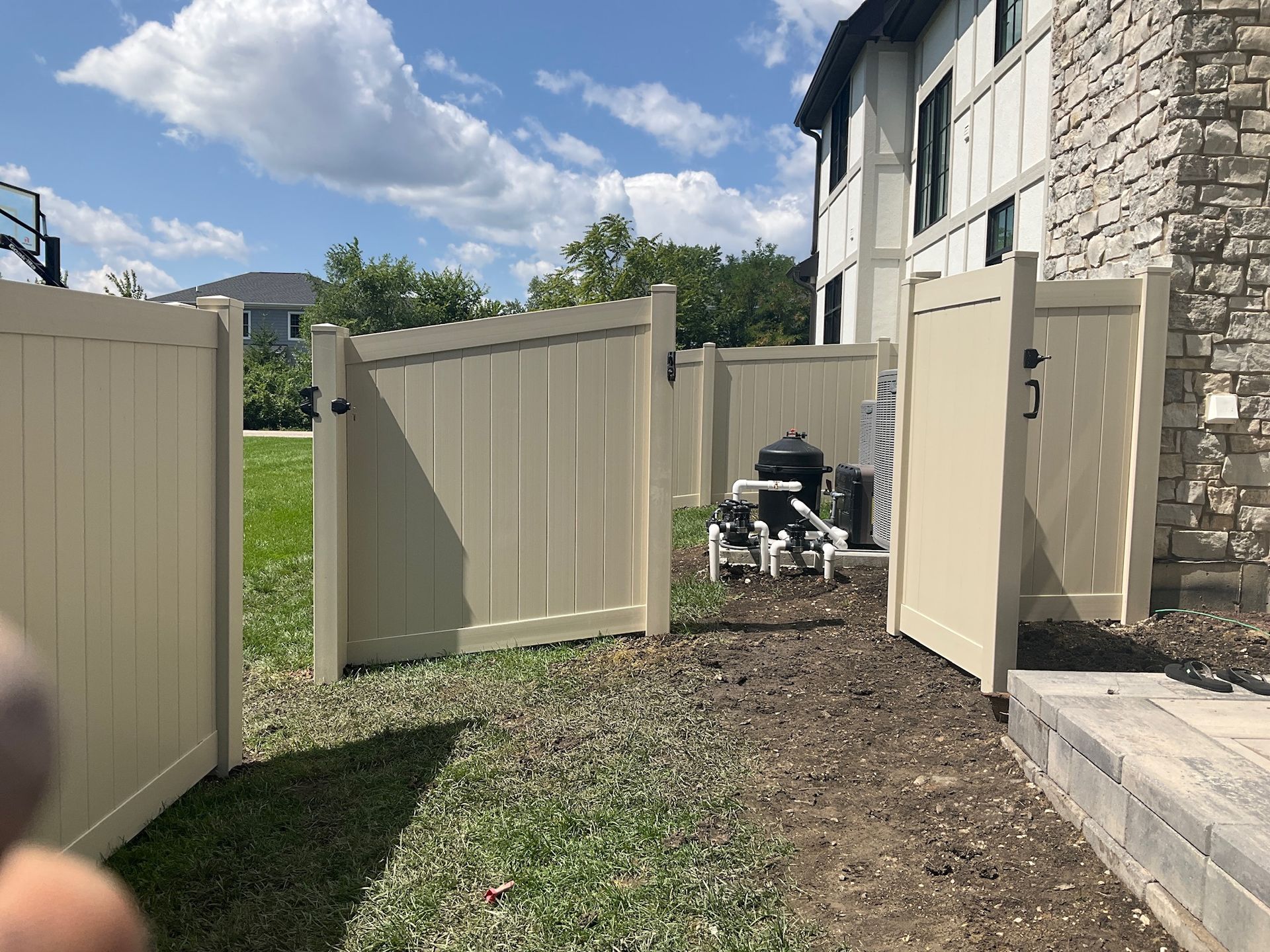 Tan vinyl fence enclosing pool equipment near a building with stone siding and a blue sky.