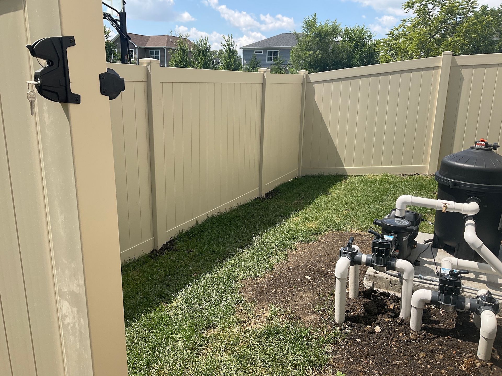 Backyard with tan vinyl fence and pool equipment. Grassy area, sunny day.