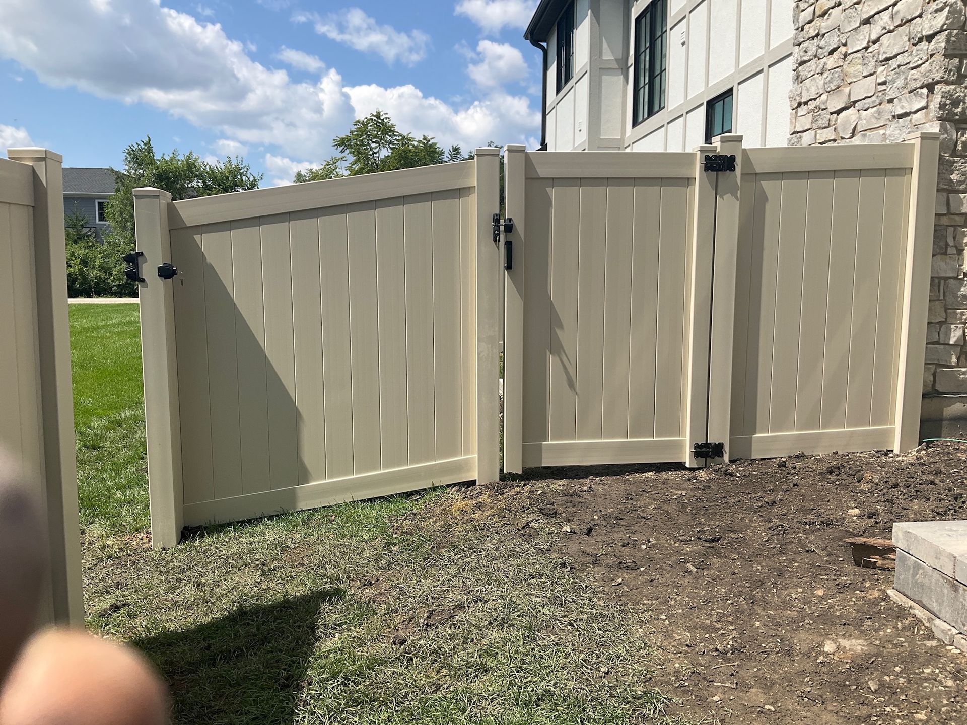 Tan vinyl fence with gate, set on grass, next to a building with stone siding.