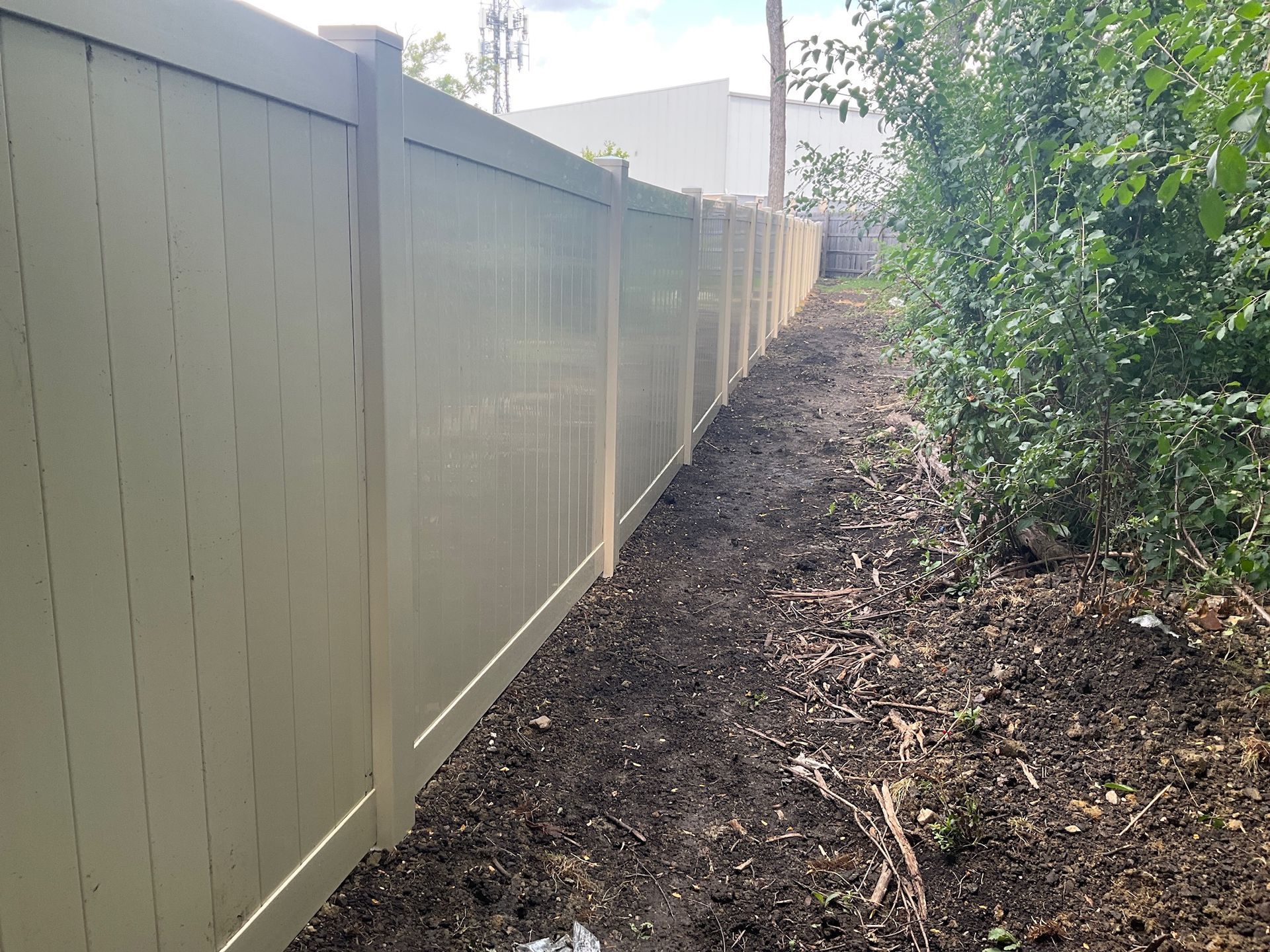 Tan vinyl fence alongside dark soil and bushes, stretching into the distance.