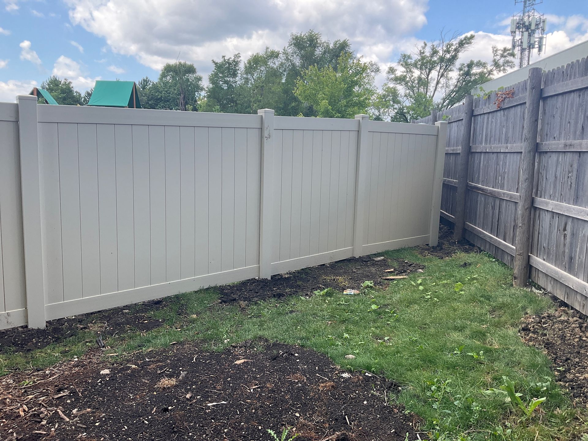 Tan vinyl fence next to a weathered wooden fence, with green grass and a blue sky in the background.