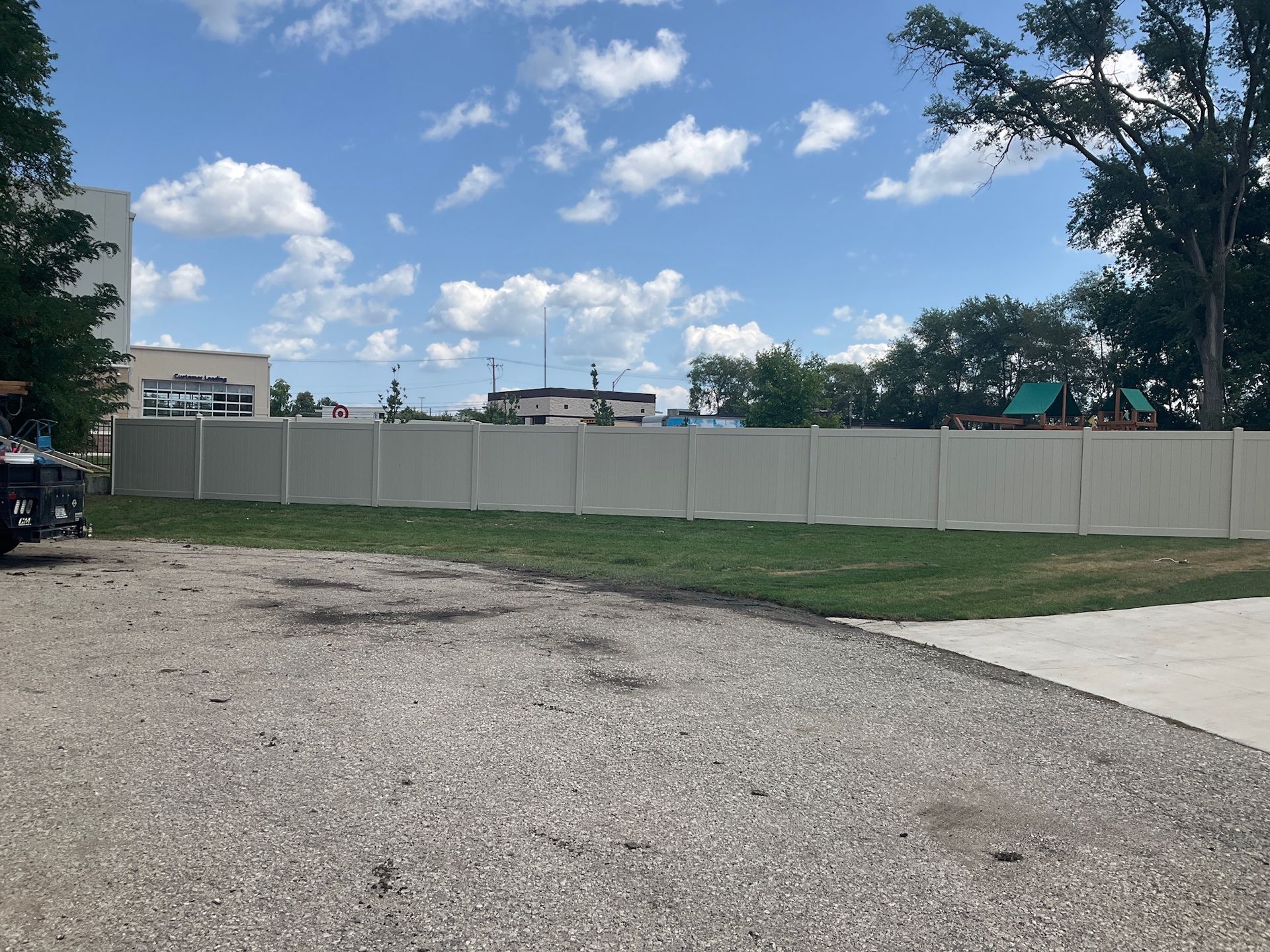 Gravel parking area in front of a long beige fence with blue sky and trees in the background.