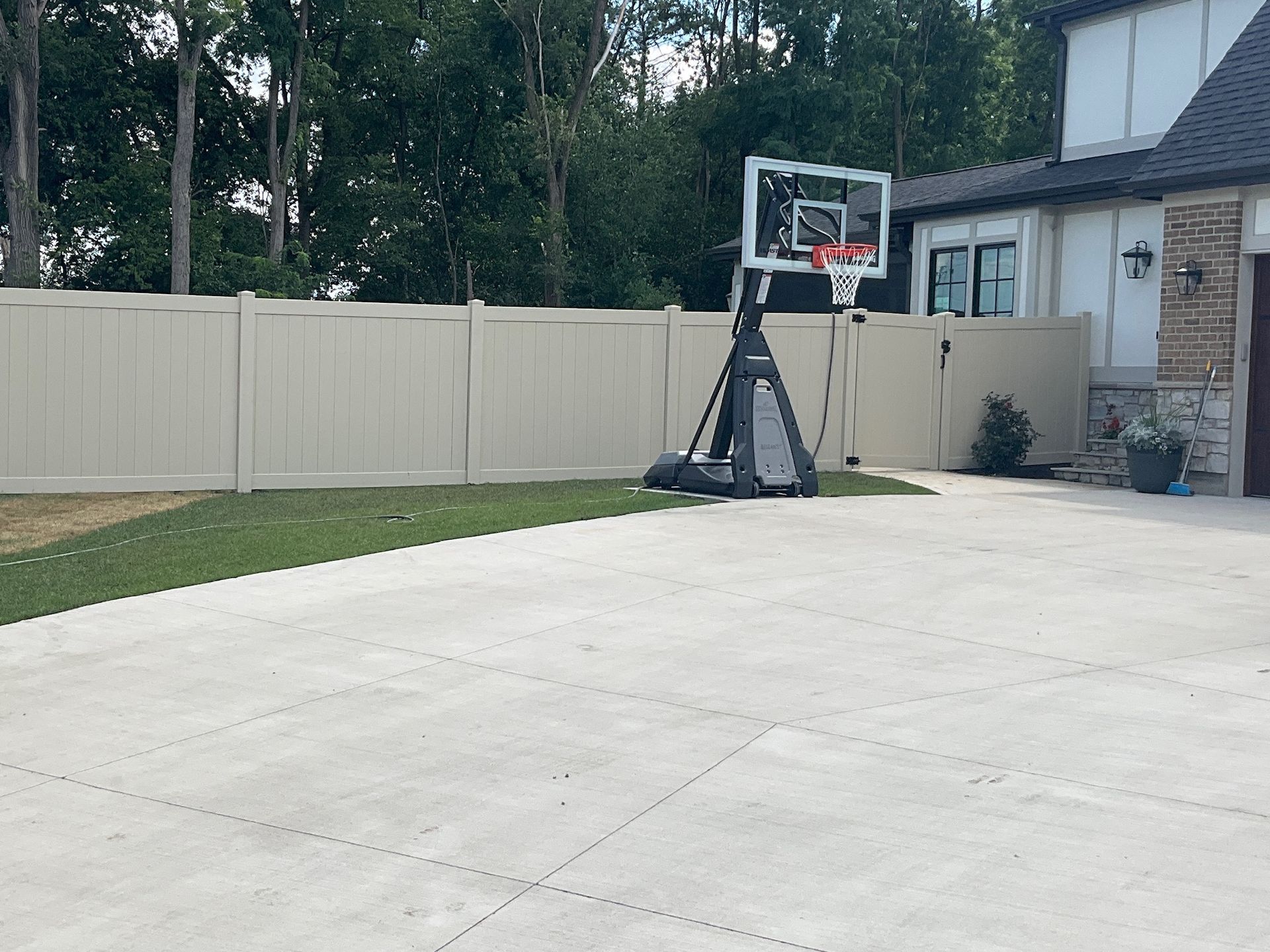 Basketball hoop on a concrete driveway in front of a house, beige fence, and trees.