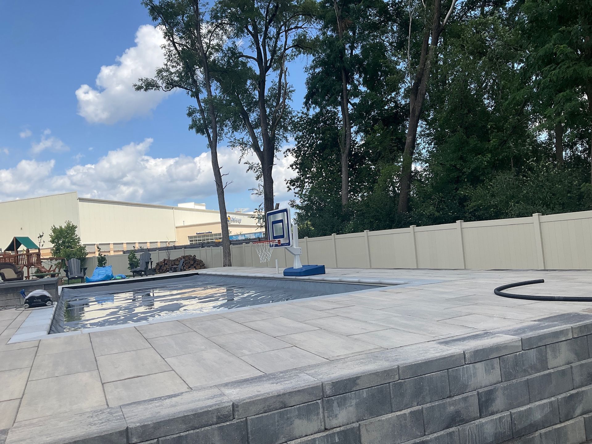 A paved patio with a basketball hoop, water, and a fence, with trees and a building in the background.