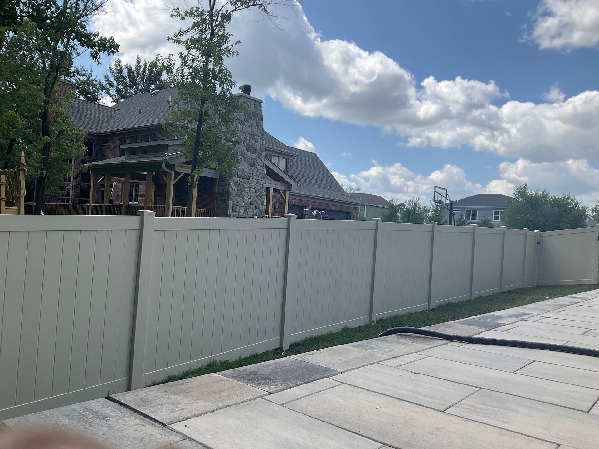 Tan vinyl fence surrounds a large house with stone facade, under a partly cloudy sky.