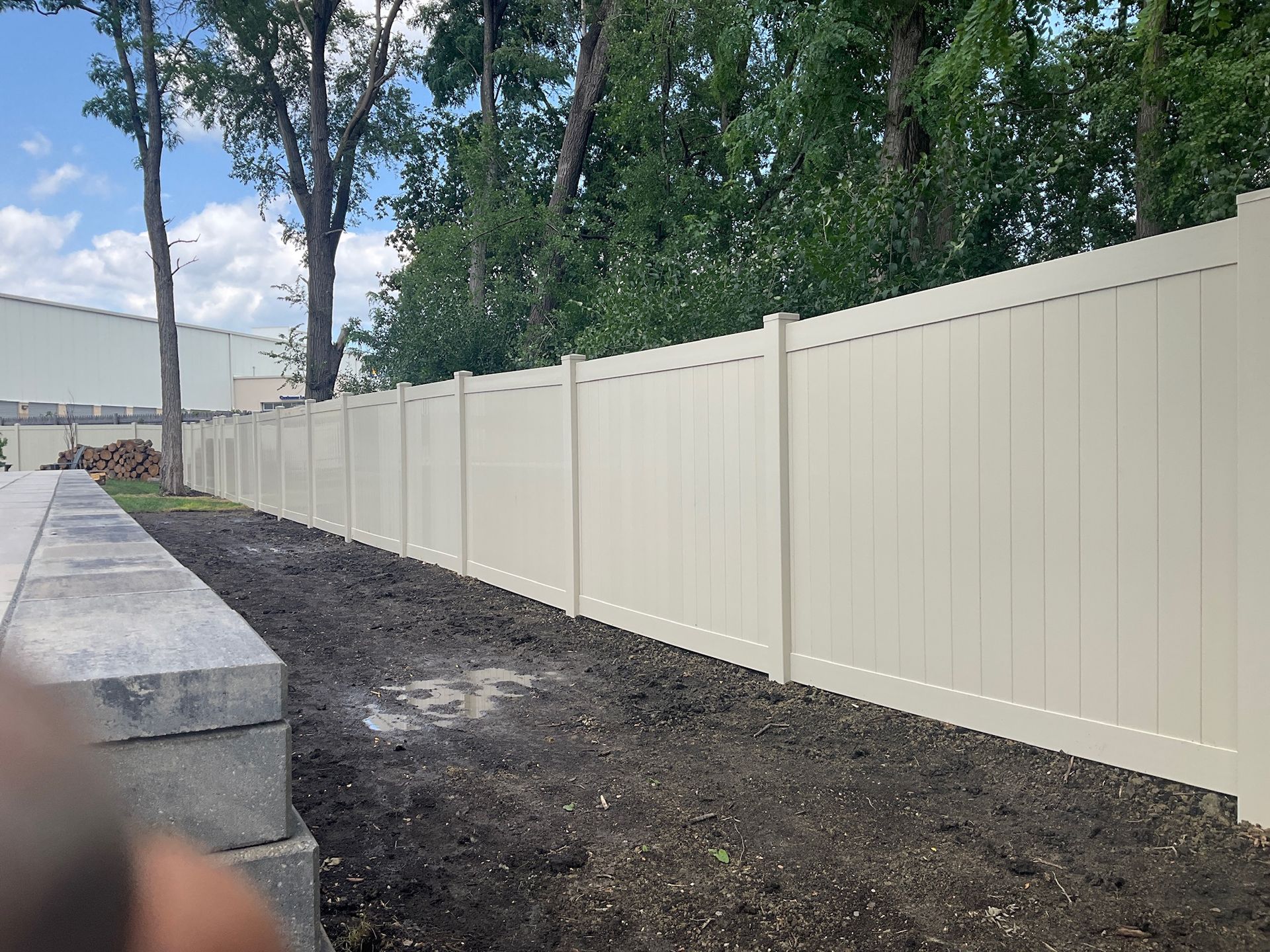 Tan vinyl fence along a dirt area with trees in the background. Gray stone blocks are visible on the left.