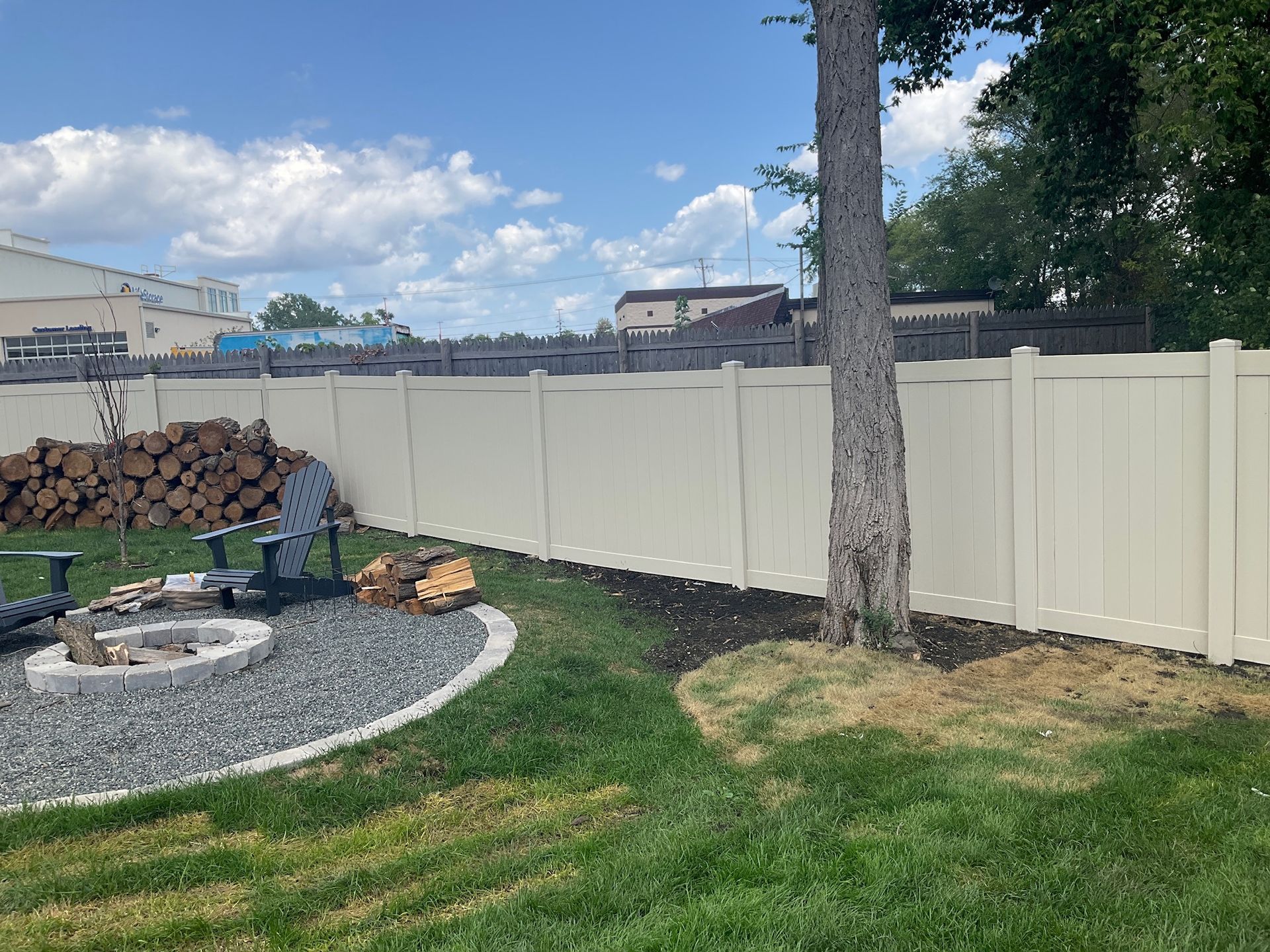 Beige vinyl fence encloses a backyard with a fire pit, firewood, and a tree. Sunny day with a blue sky.