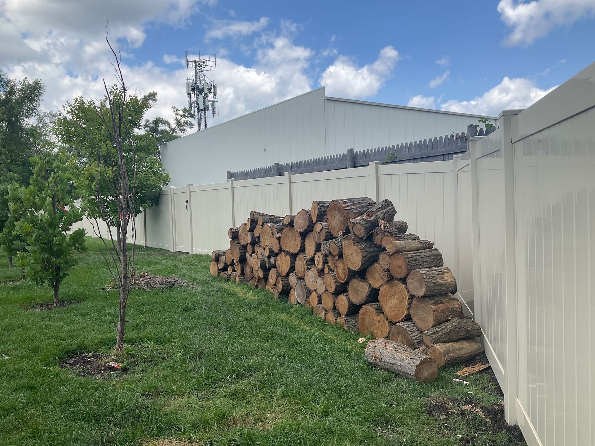 Pile of firewood stacked on green grass next to a white fence and building under a blue sky.