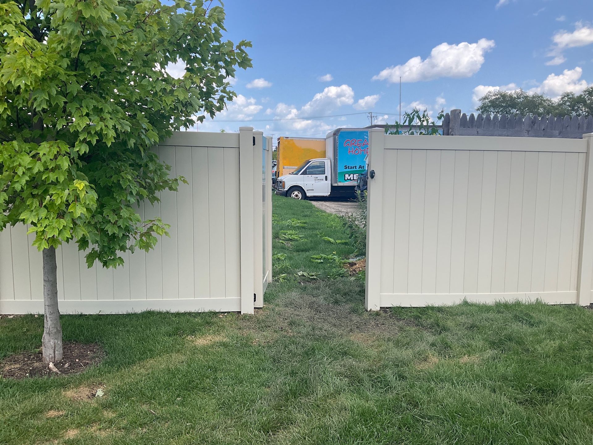 Open beige gate in a fenced yard, revealing a glimpse of trucks on a sunny day.
