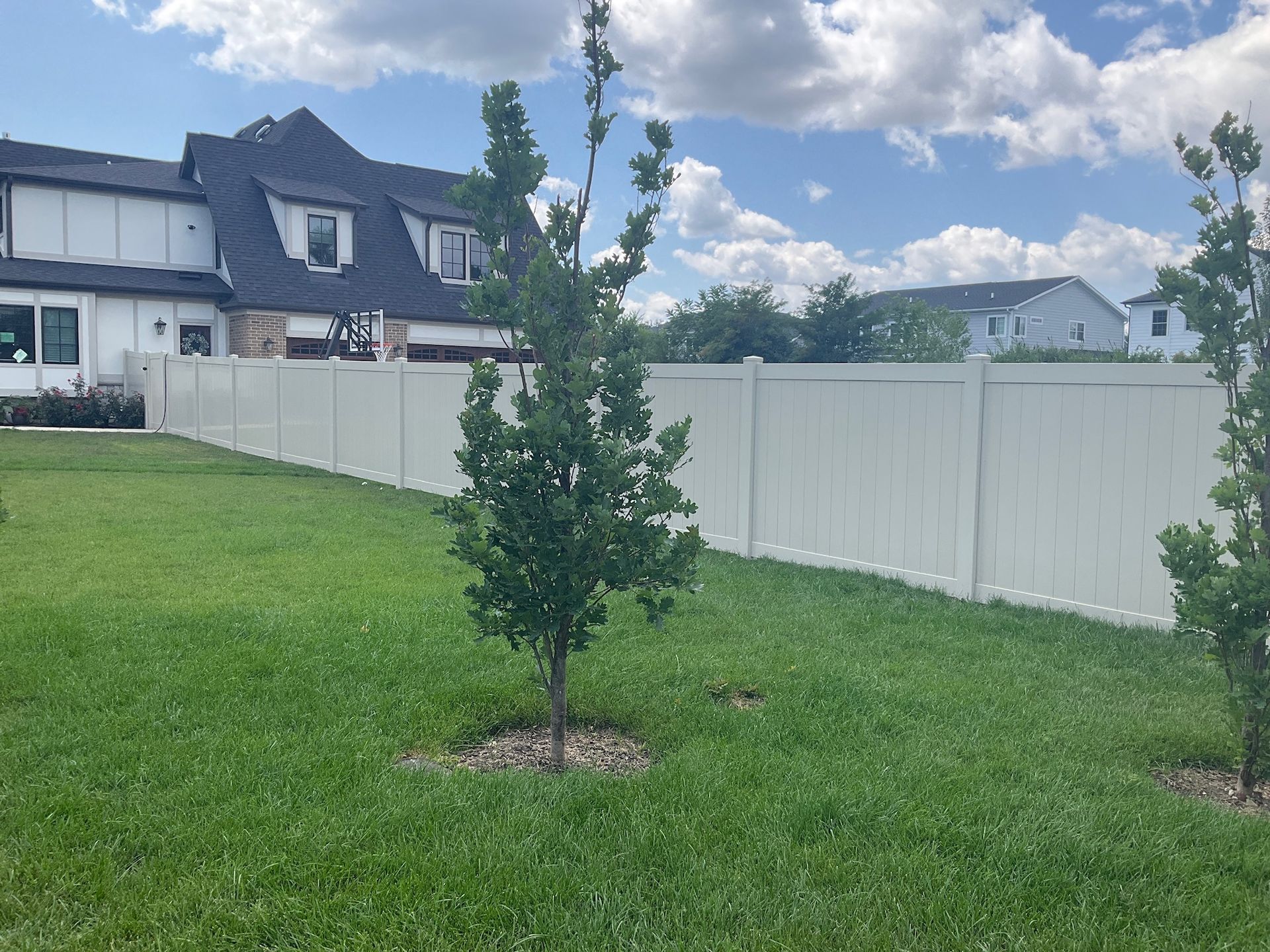 White vinyl fence in a grassy yard, with a tree in front of it and a house in the background on a partly cloudy day.