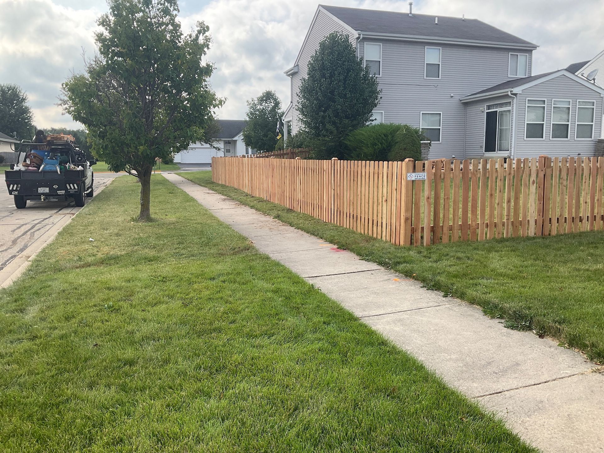 A newly built wooden fence lines the front yard of a two-story house, next to a sidewalk and green lawn.