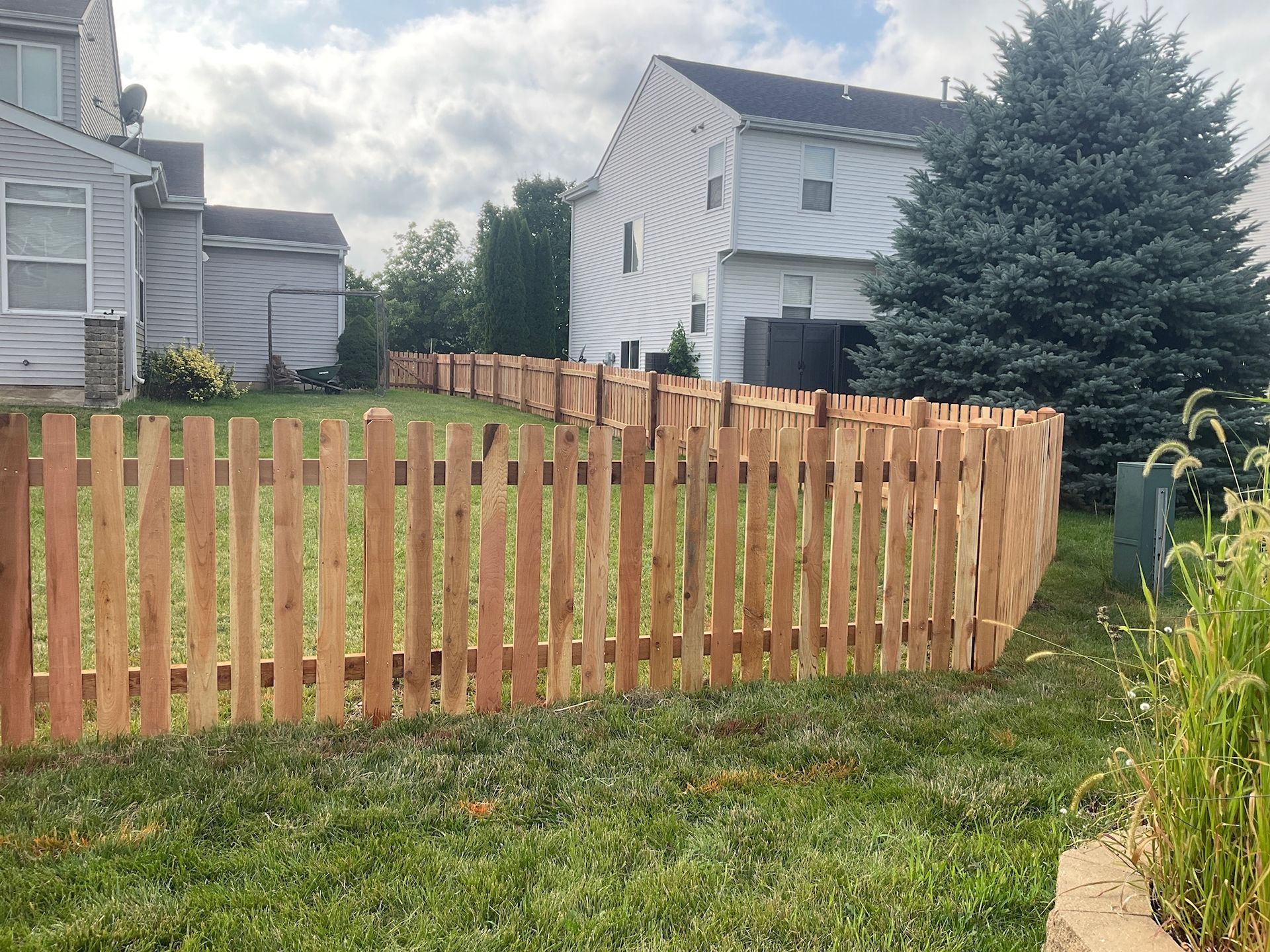 Wooden picket fence in a grassy backyard, curving toward houses under a cloudy sky.