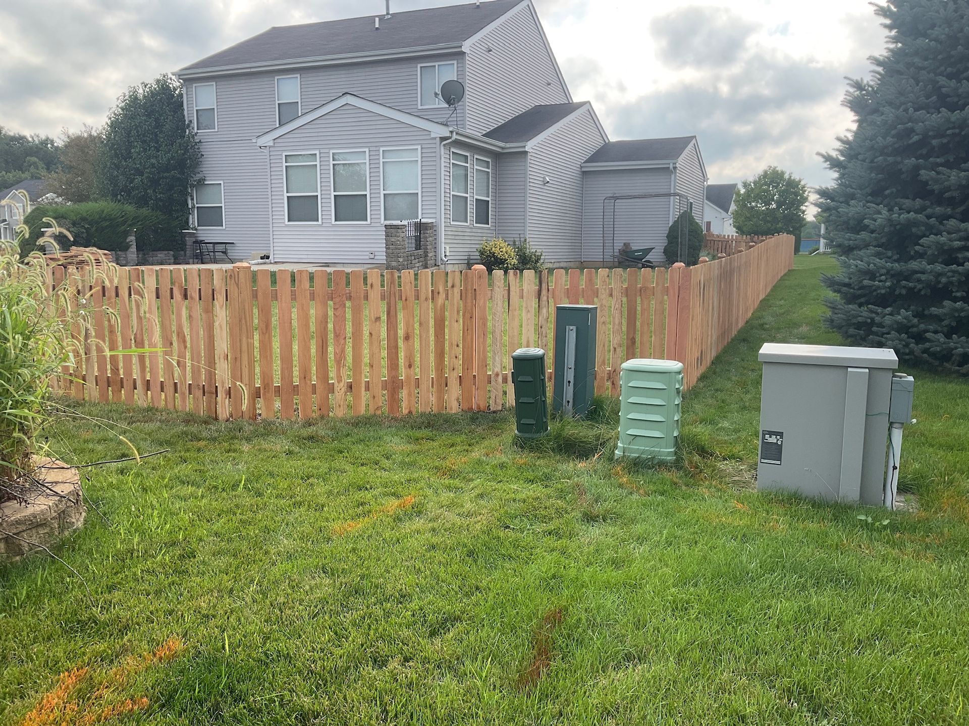 Wooden fence along a grassy backyard, with utility boxes and a house in the background.