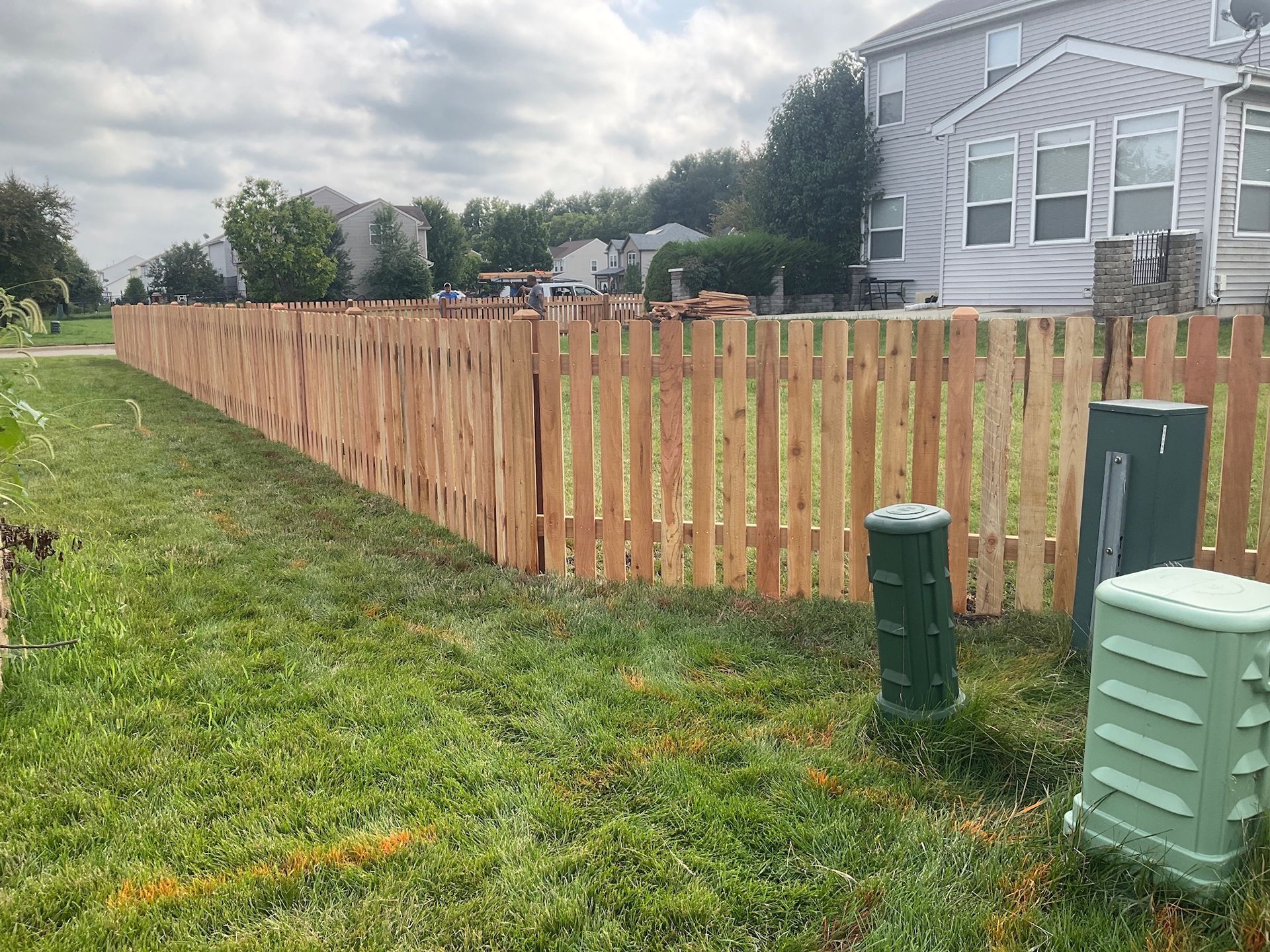 A wooden fence with green utility boxes next to it, bordering a grassy area and houses.
