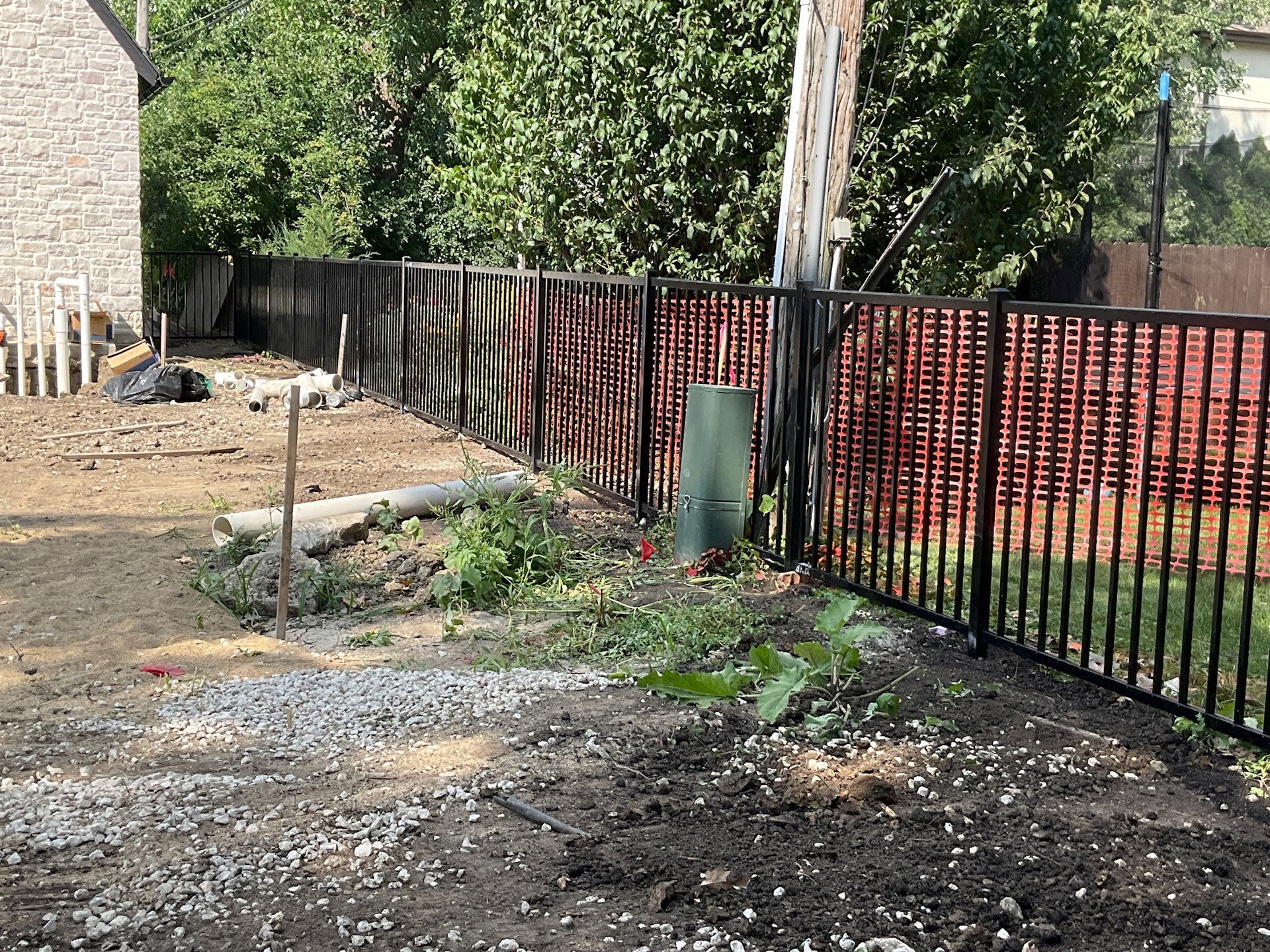 Black metal fence bordering a yard with dirt, gravel, and construction debris. Green plants and trees in the background.