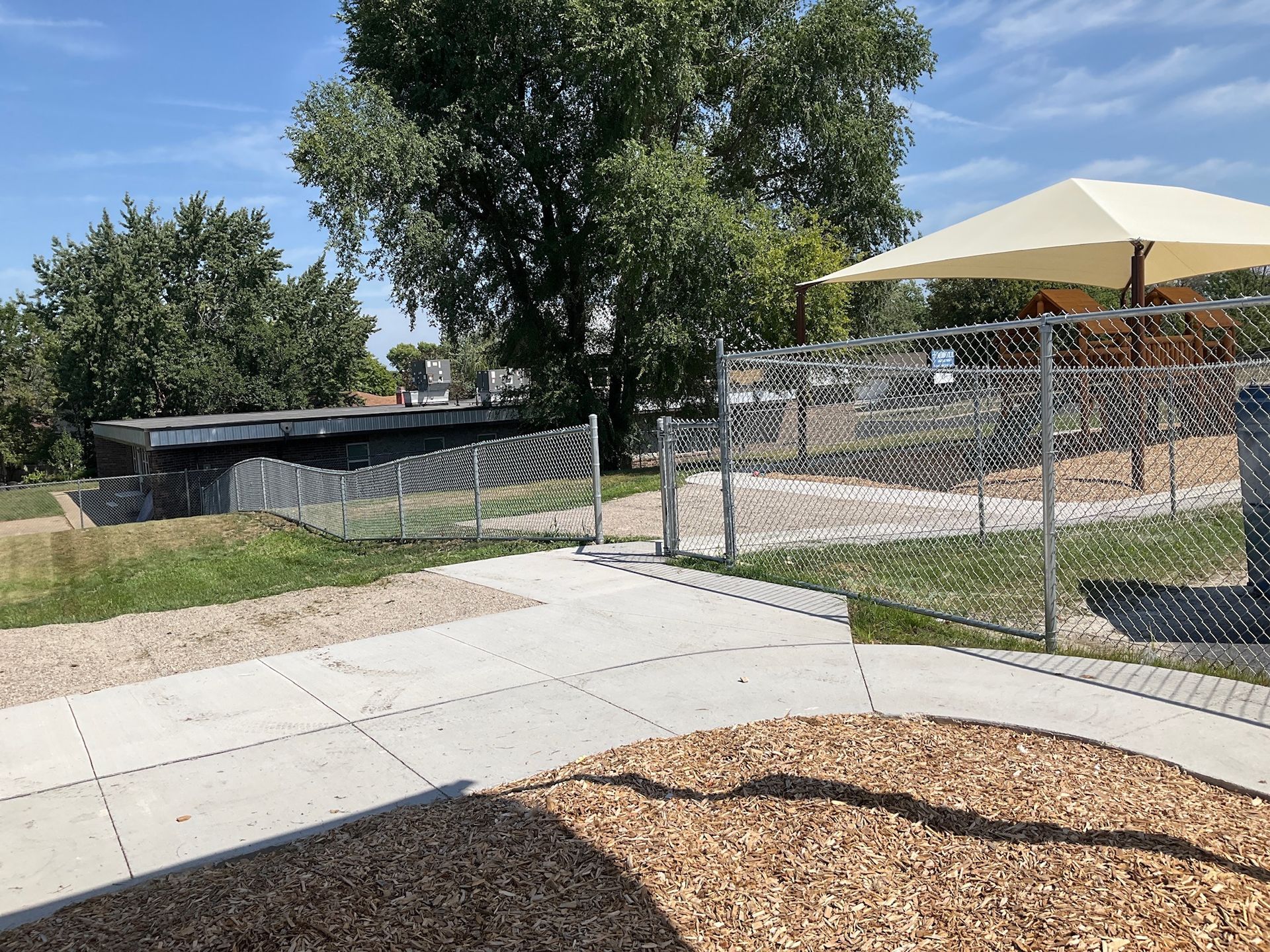 A playground entrance with a concrete path, wood chips, and a chain-link fence on a sunny day.