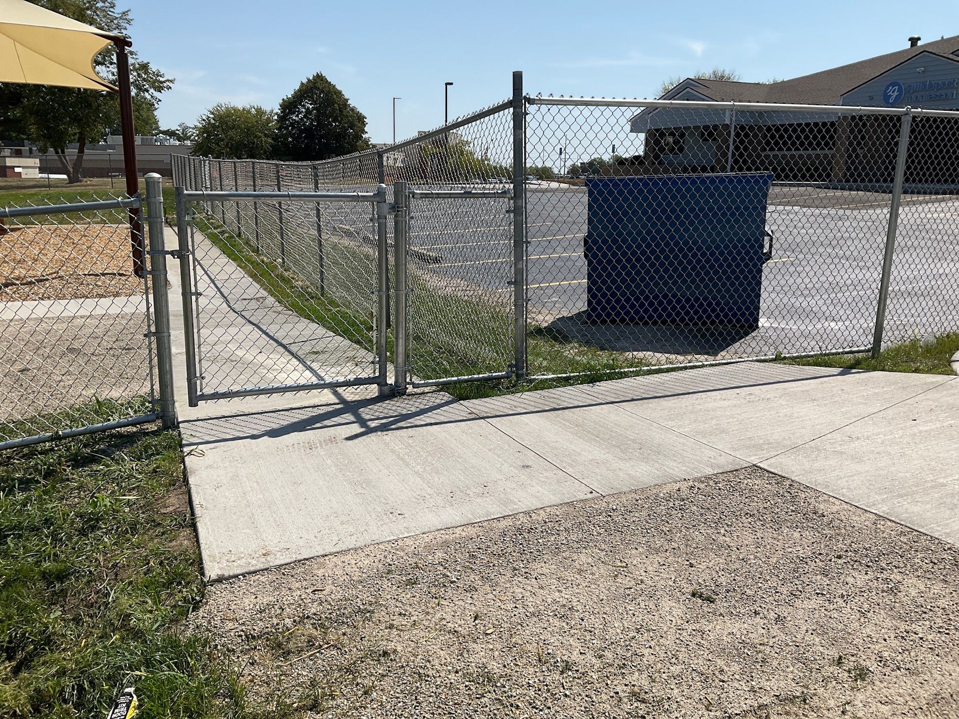 Chain-link fence with gate, concrete walkway, and dumpster in a paved lot.