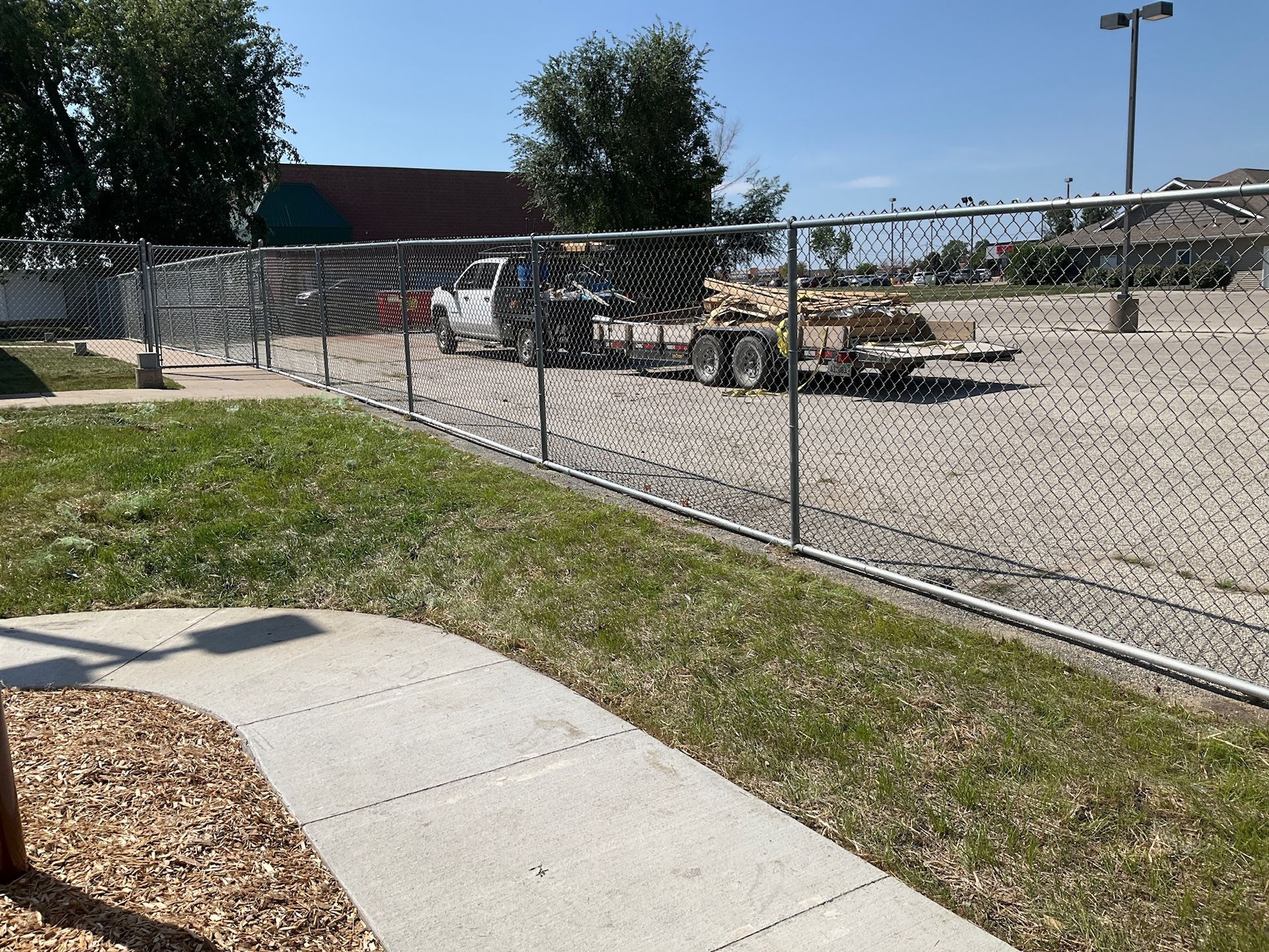 Chain-link fence encloses a gravel lot with construction equipment. Green grass and a sidewalk are in the foreground.
