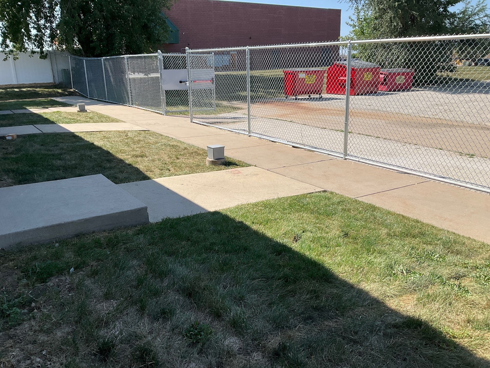 Chain link fence along a concrete path and grass, leading to a red container building.
