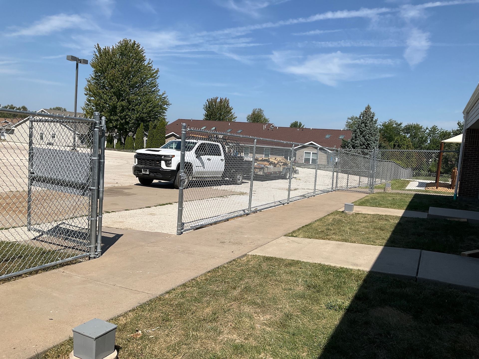 White truck inside a chain-link fence, gate open on a paved walkway, grass and buildings in background, blue sky.