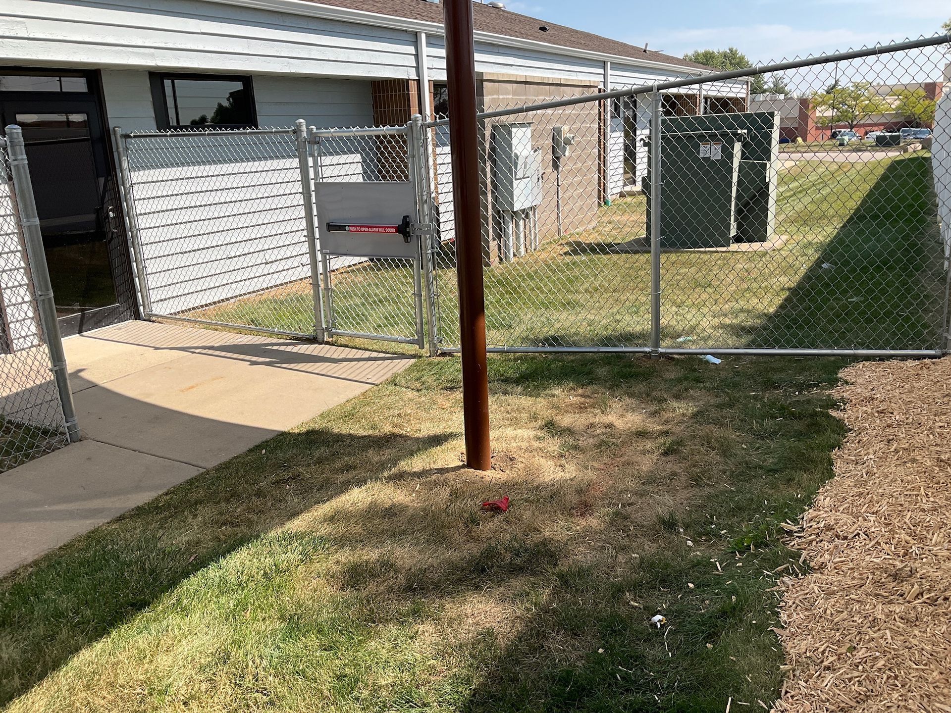 Chain-link fence encloses an electrical area with a pole in the center. Green and brown grass in the yard.