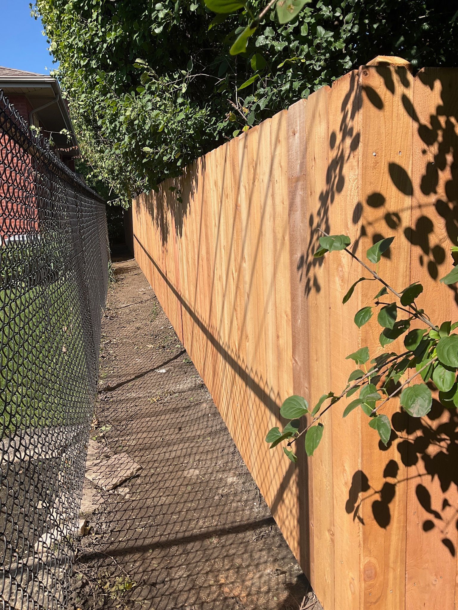 Wooden fence and camouflage netting along a narrow path, trees overhead.