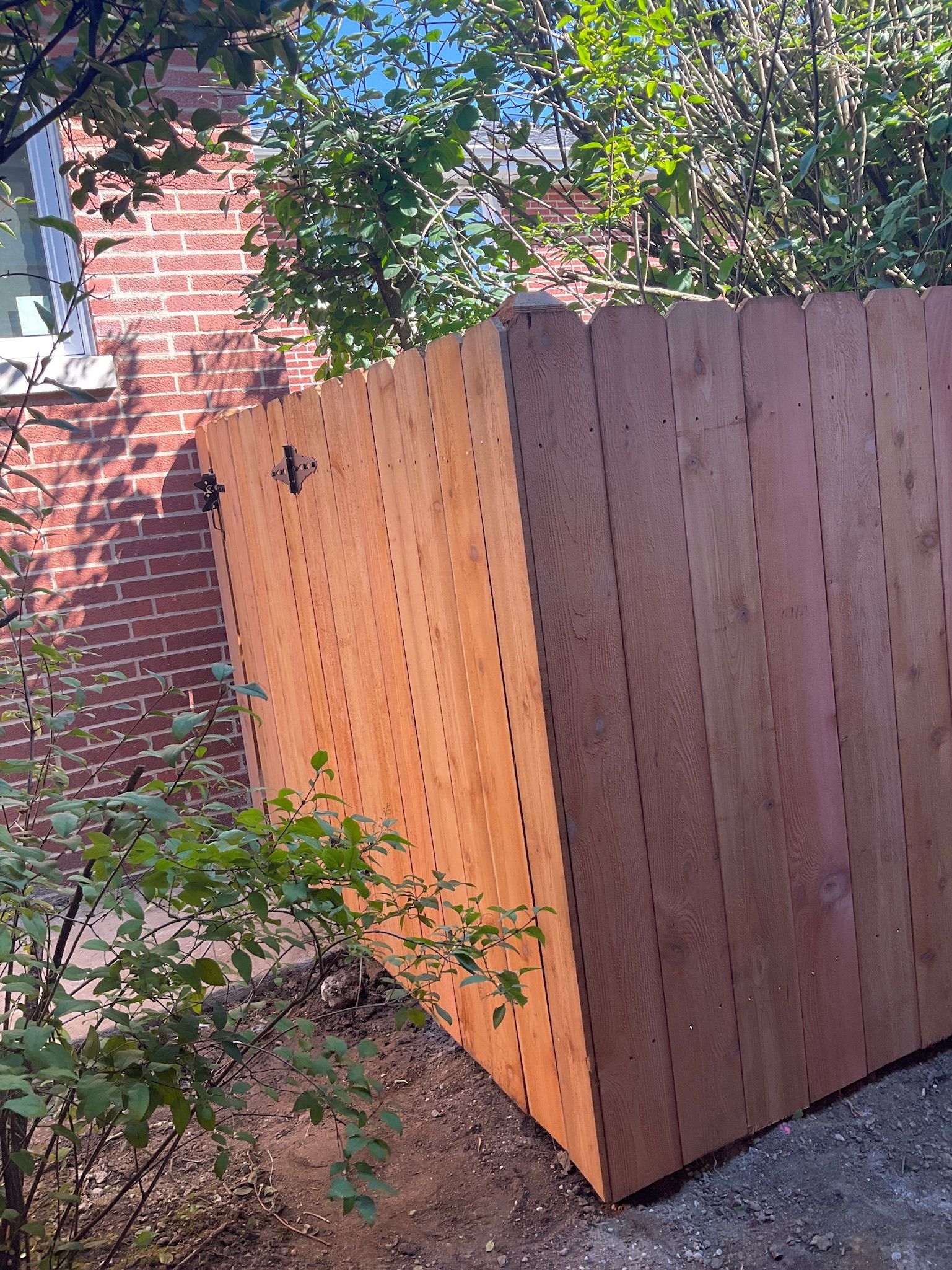 Wooden fence leaning, possibly damaged, near brick building and greenery.