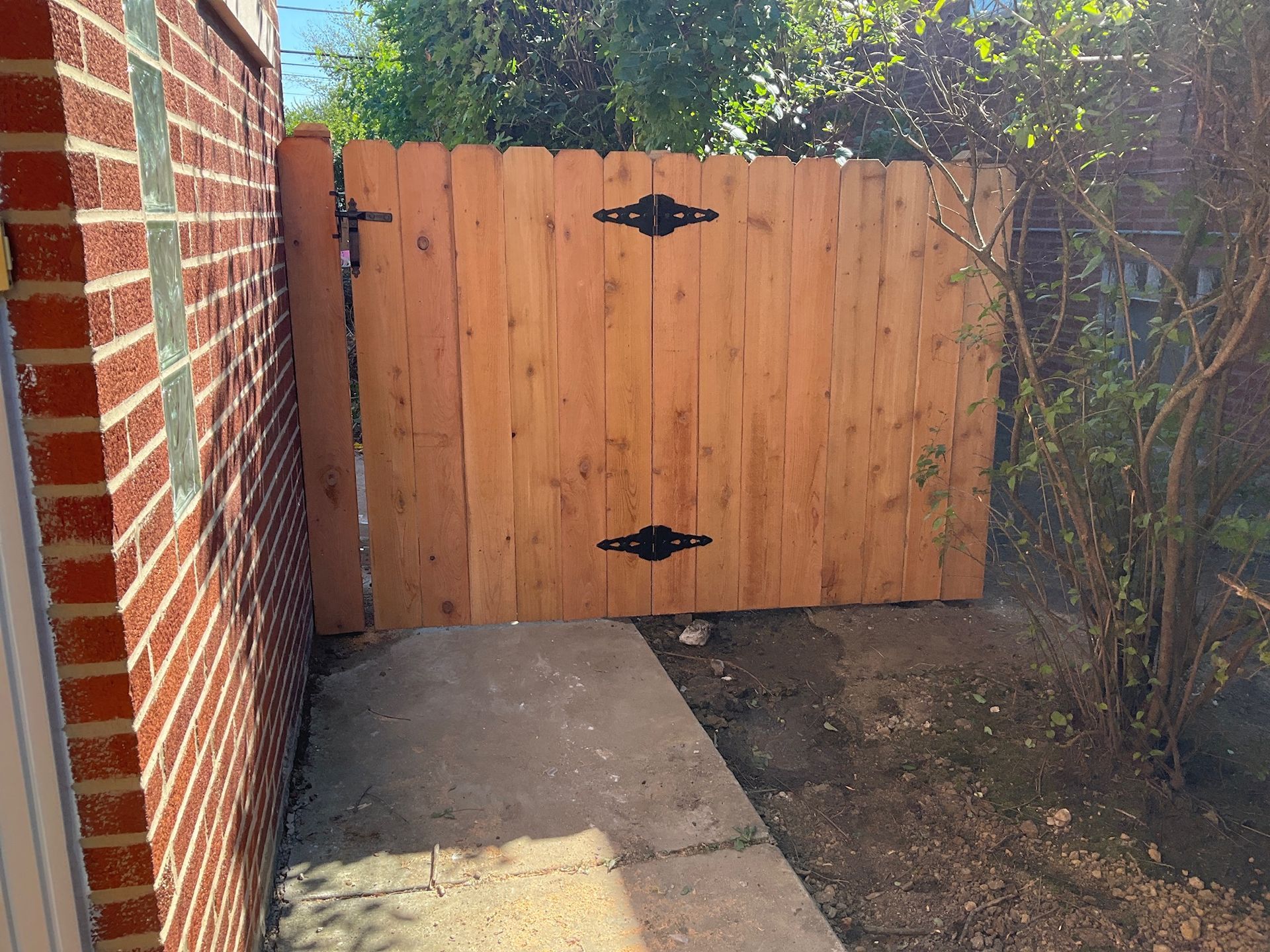Wooden gate with black hinges and latch, between a brick building and shrubbery.