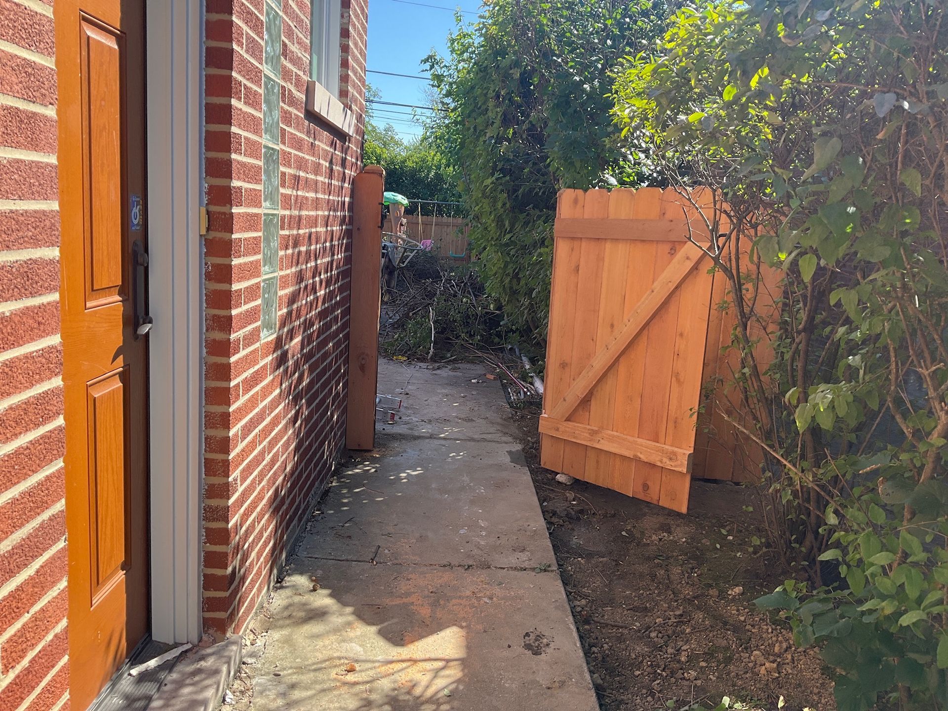 Narrow walkway beside a red brick wall, leading toward a wooden gate; sunlit outdoor setting.