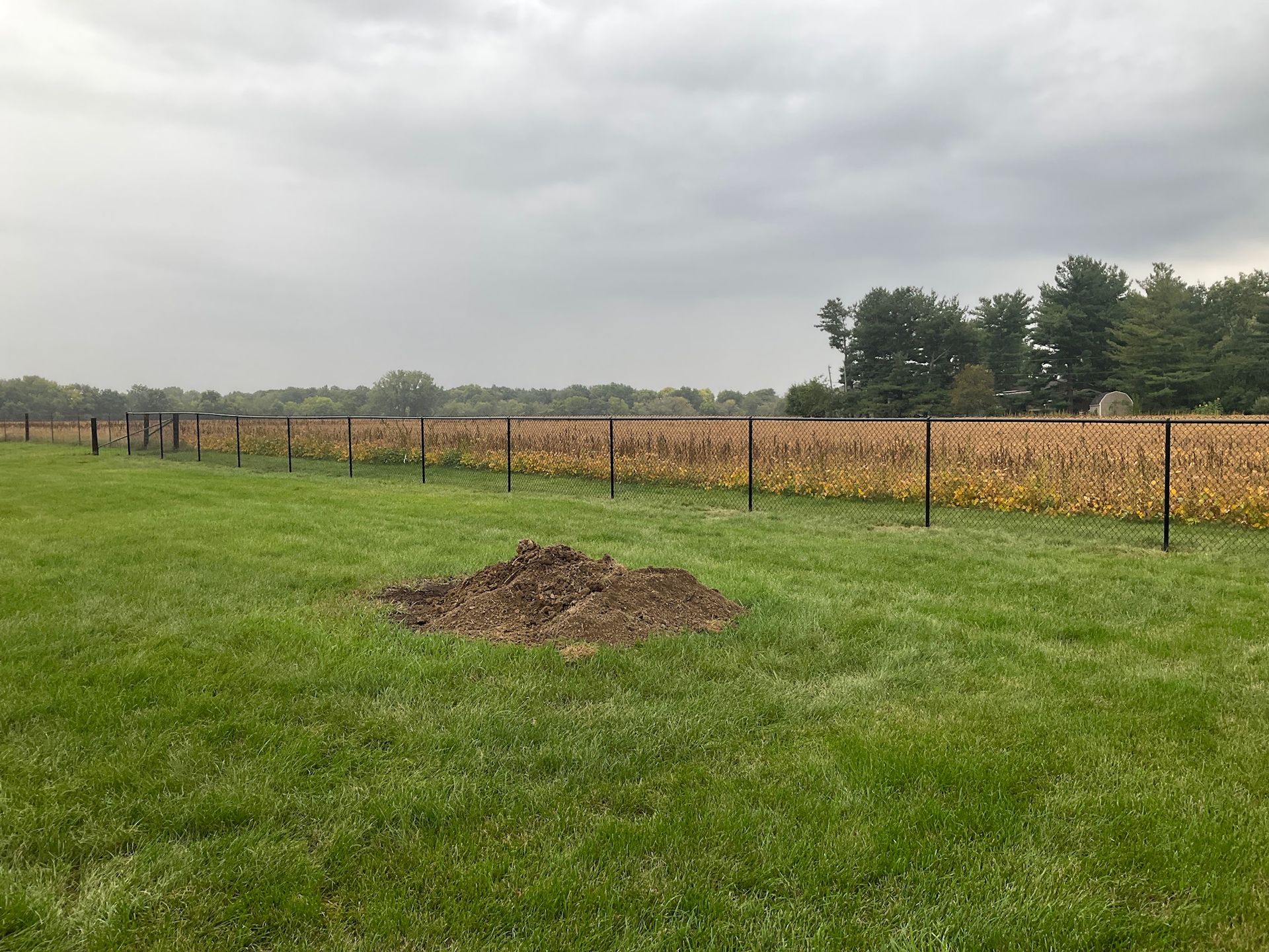 Black fence in a green field with a pile of dirt in the foreground and a crop field in the background under a cloudy sky.