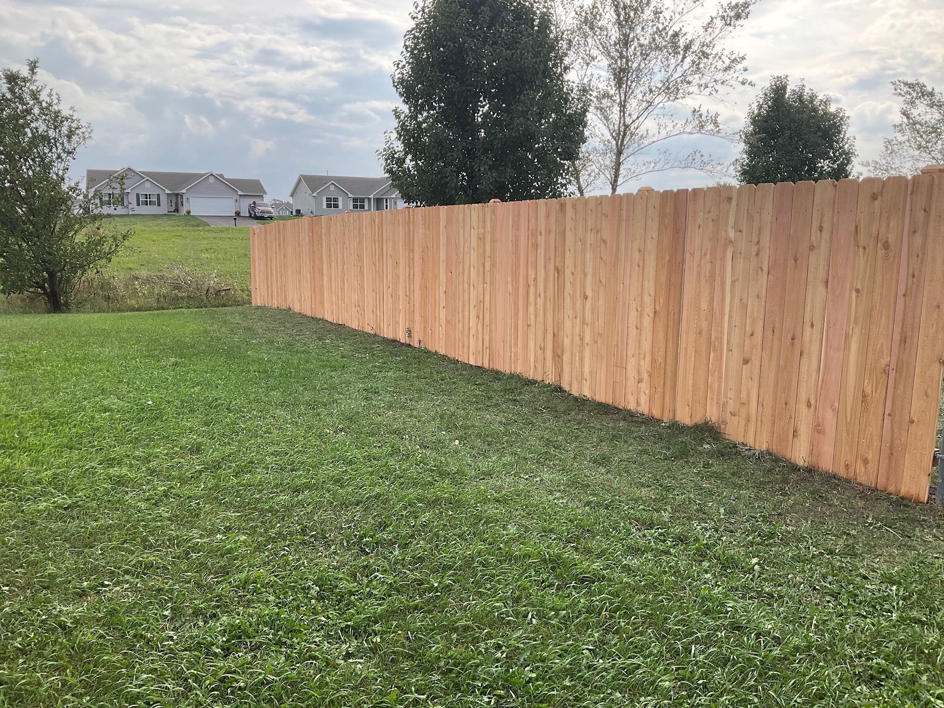 Wooden fence in grassy backyard, homes in the background under a cloudy sky.