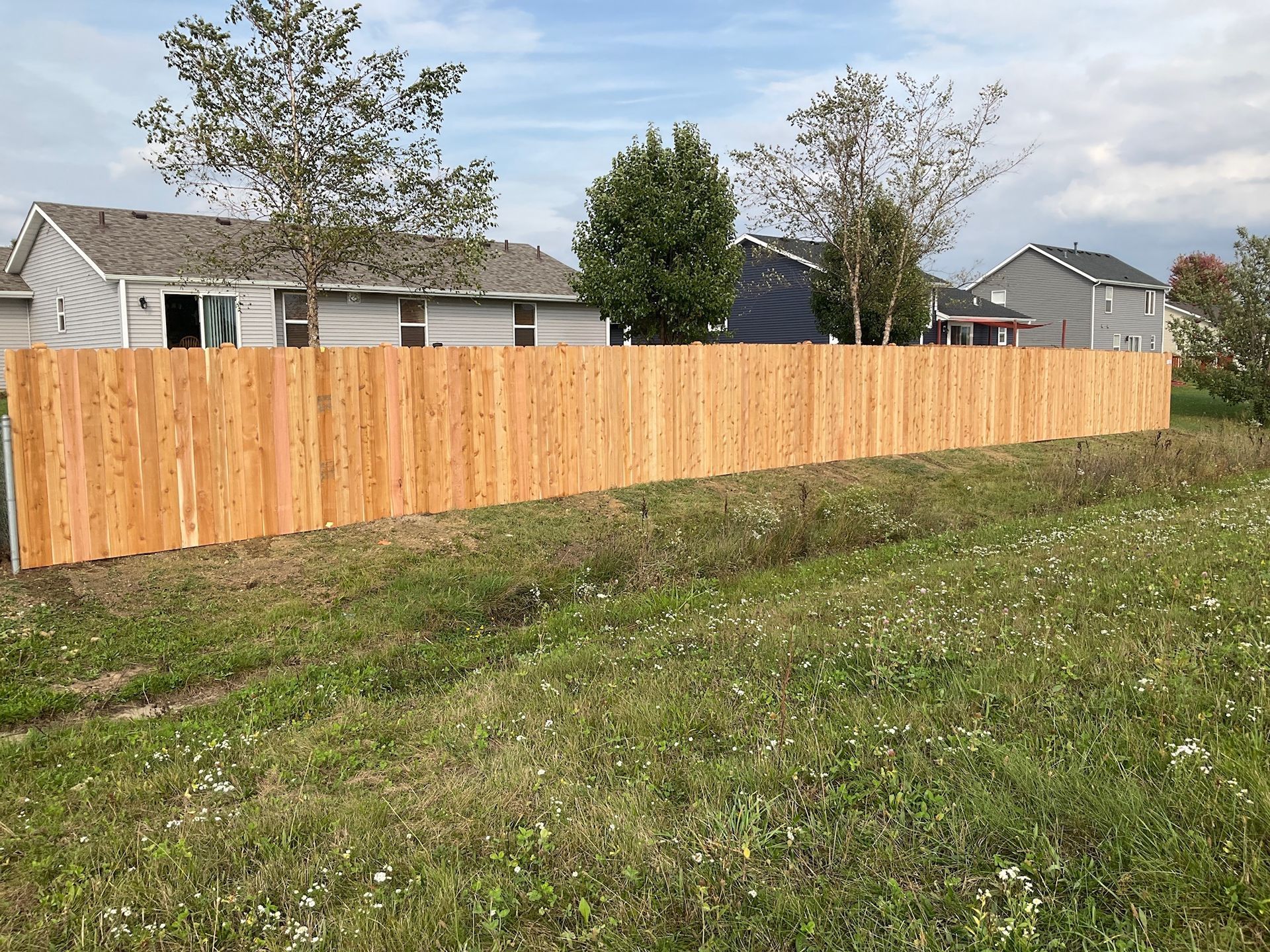 Wooden privacy fence bordering a grassy field, houses in the background under a cloudy sky.