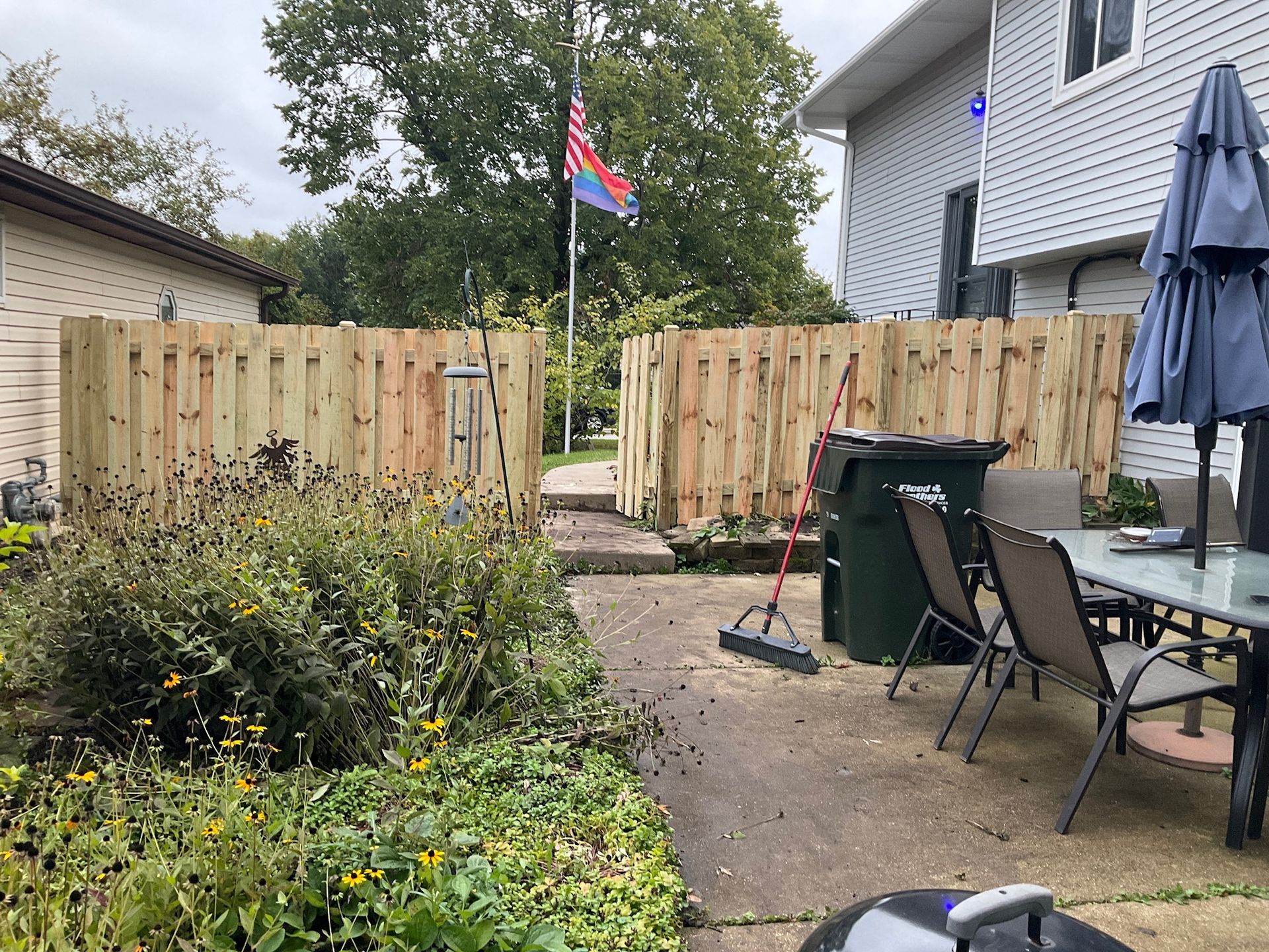 Backyard with a wooden fence, flag, patio, and green trash can.