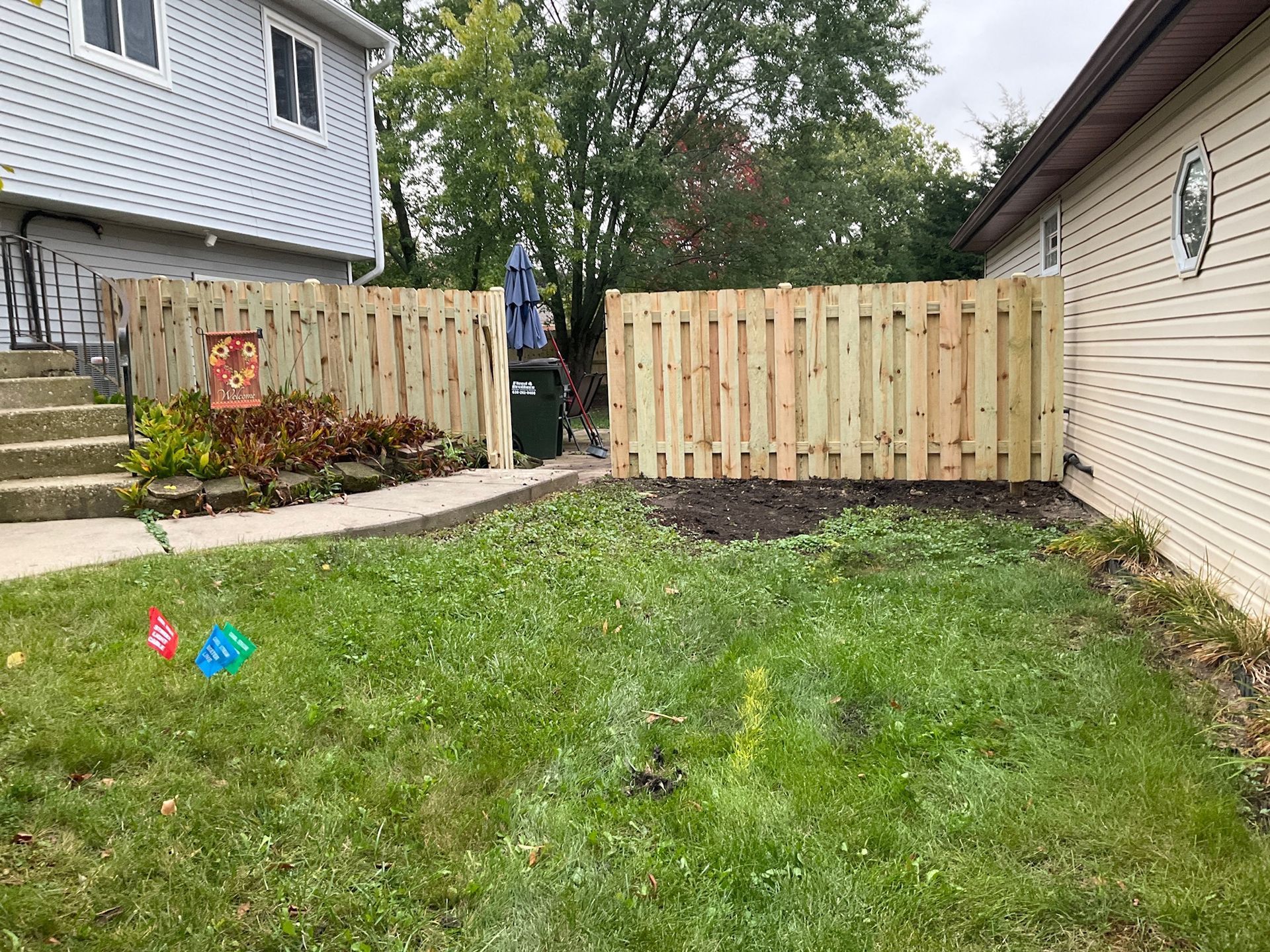 Wooden fence encloses a backyard with grass, a planting bed, and a building on each side.